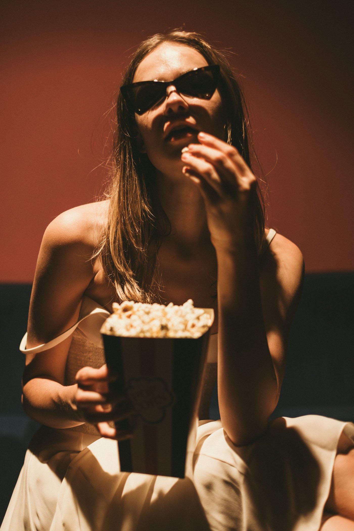 Mujer con gafas de sol oscuras, cabello largo, en un entorno con iluminación tenue, comiendo palomitas de maíz de un recipiente grande, con expresión de disfrute.