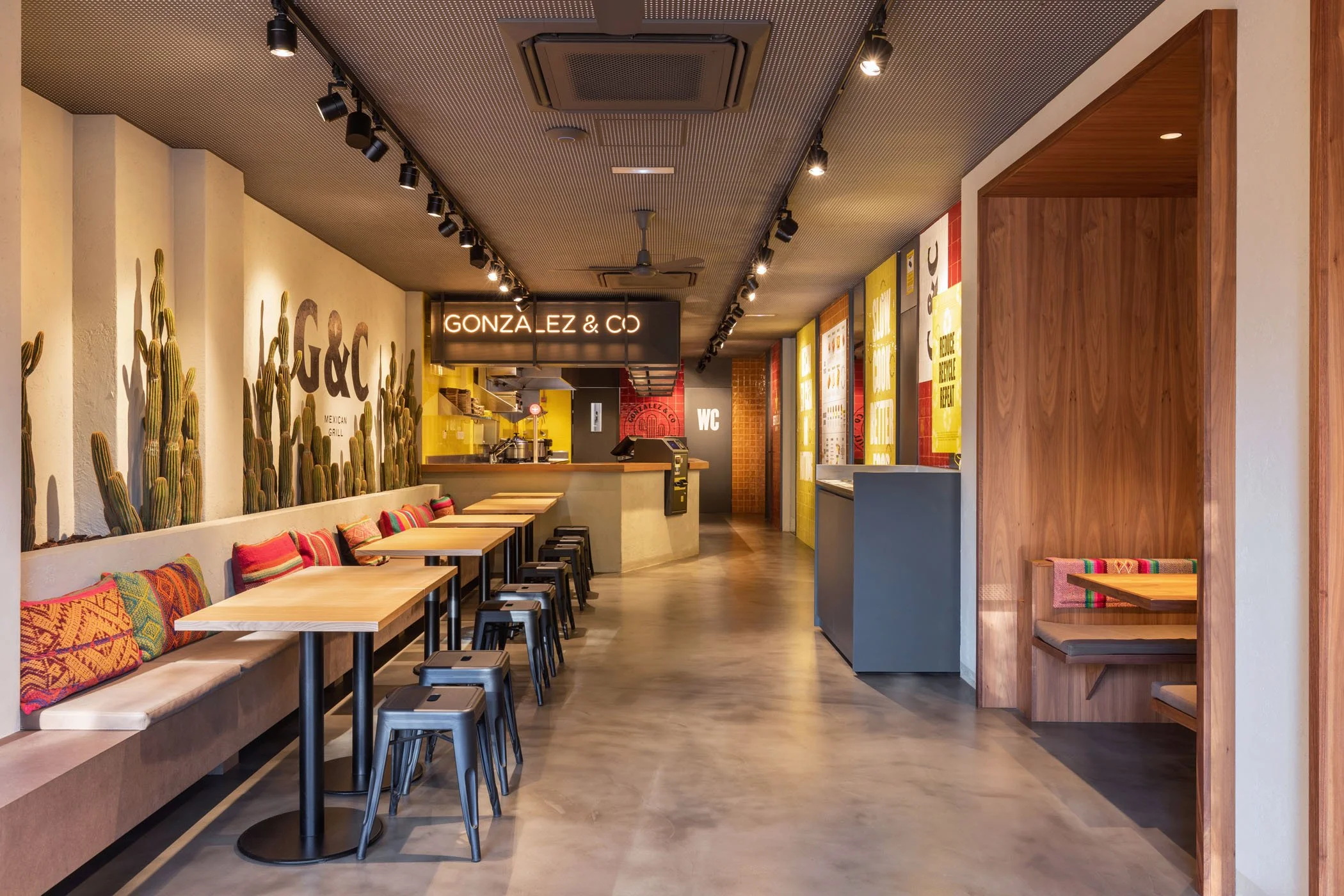 Interior de un restaurante mexicano con decoración colorida, paredes con cactus y carteles con menú, mesas de madera y bancos con cojines, área de servicio en el fondo.