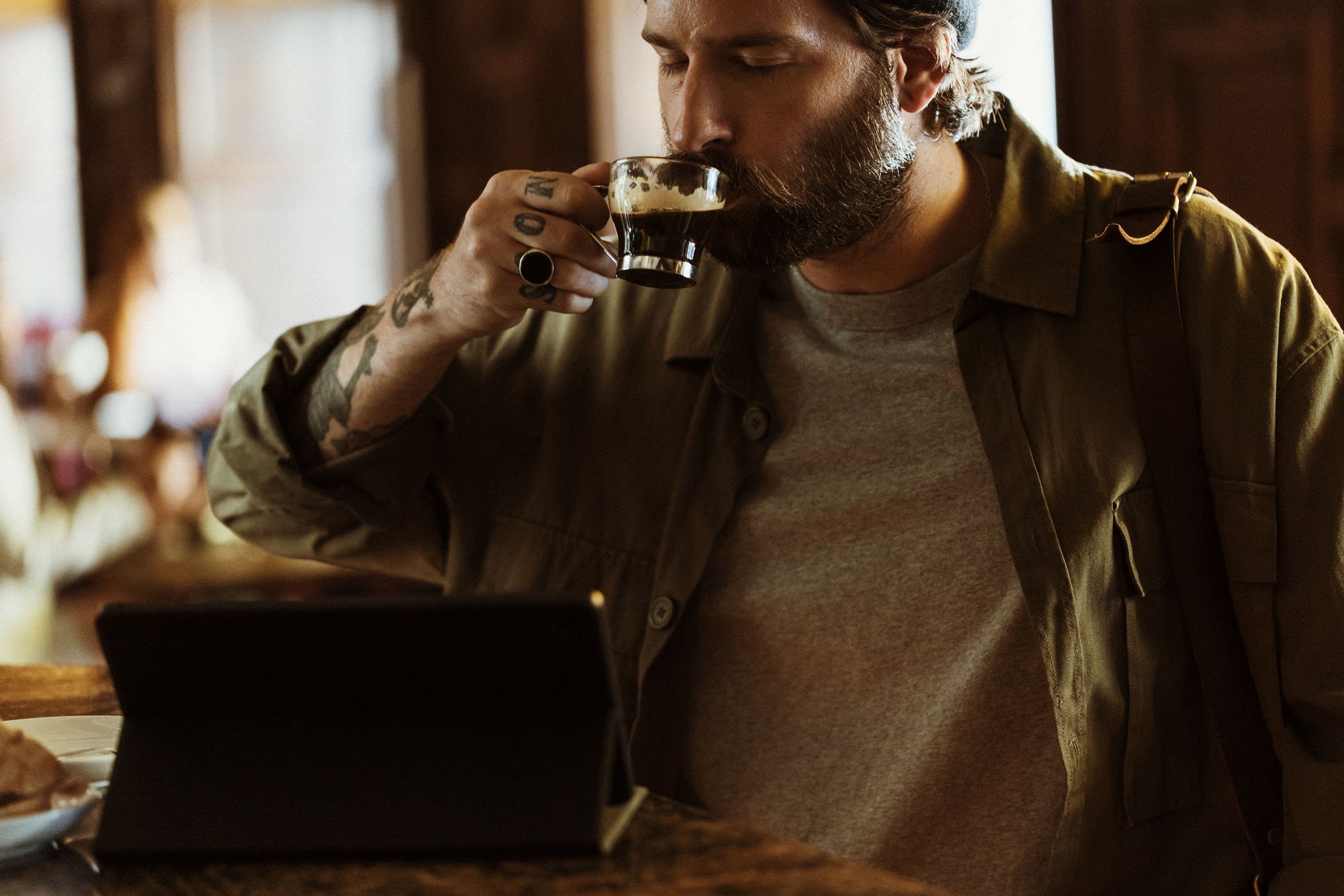 Un hombre con barba y tatuajes tomando café en un ambiente de cafetería.