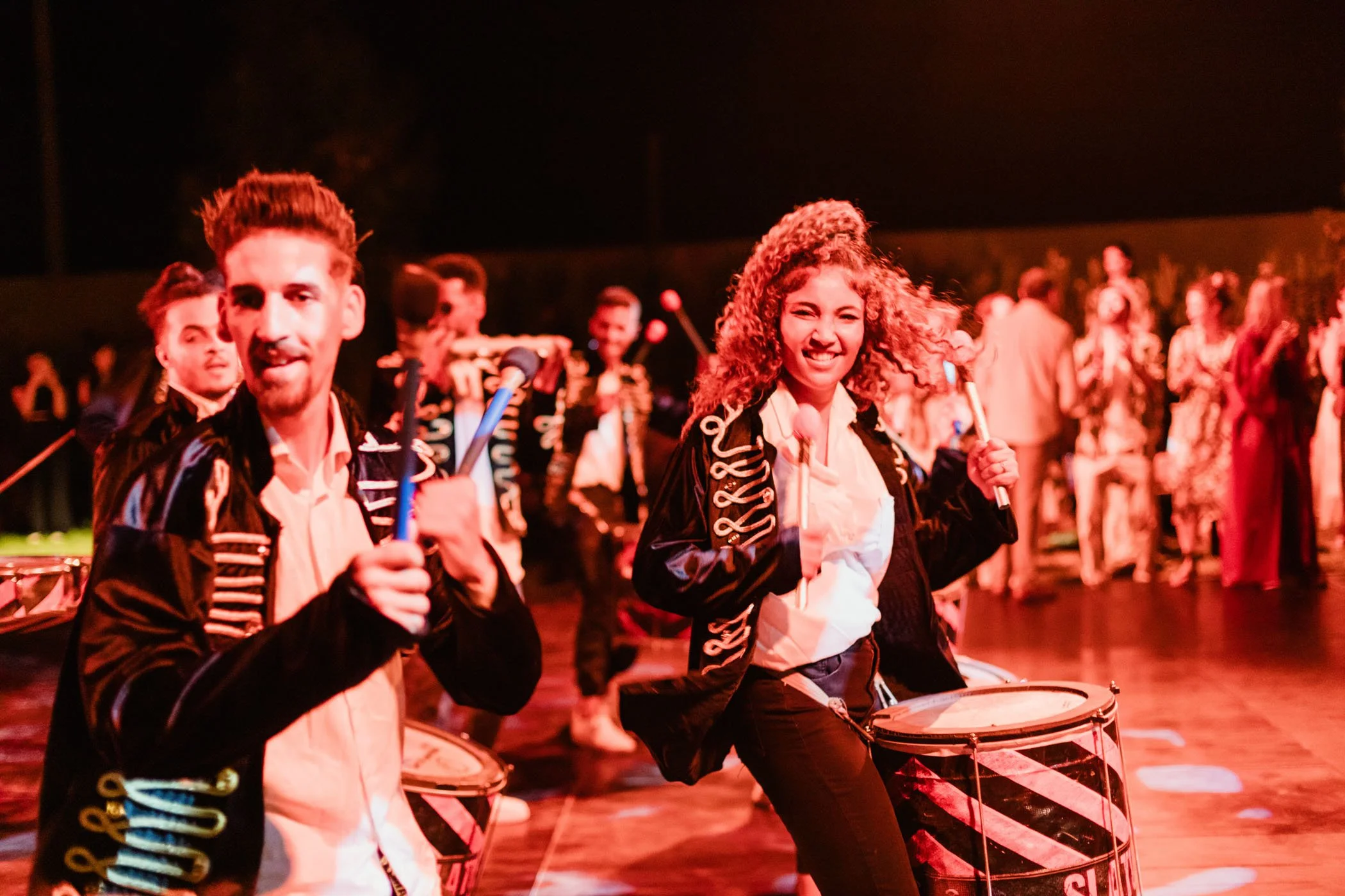 Grupo de músicos tocando batería en un escenario durante una presentación en la noche, con varios músicos de fondo.