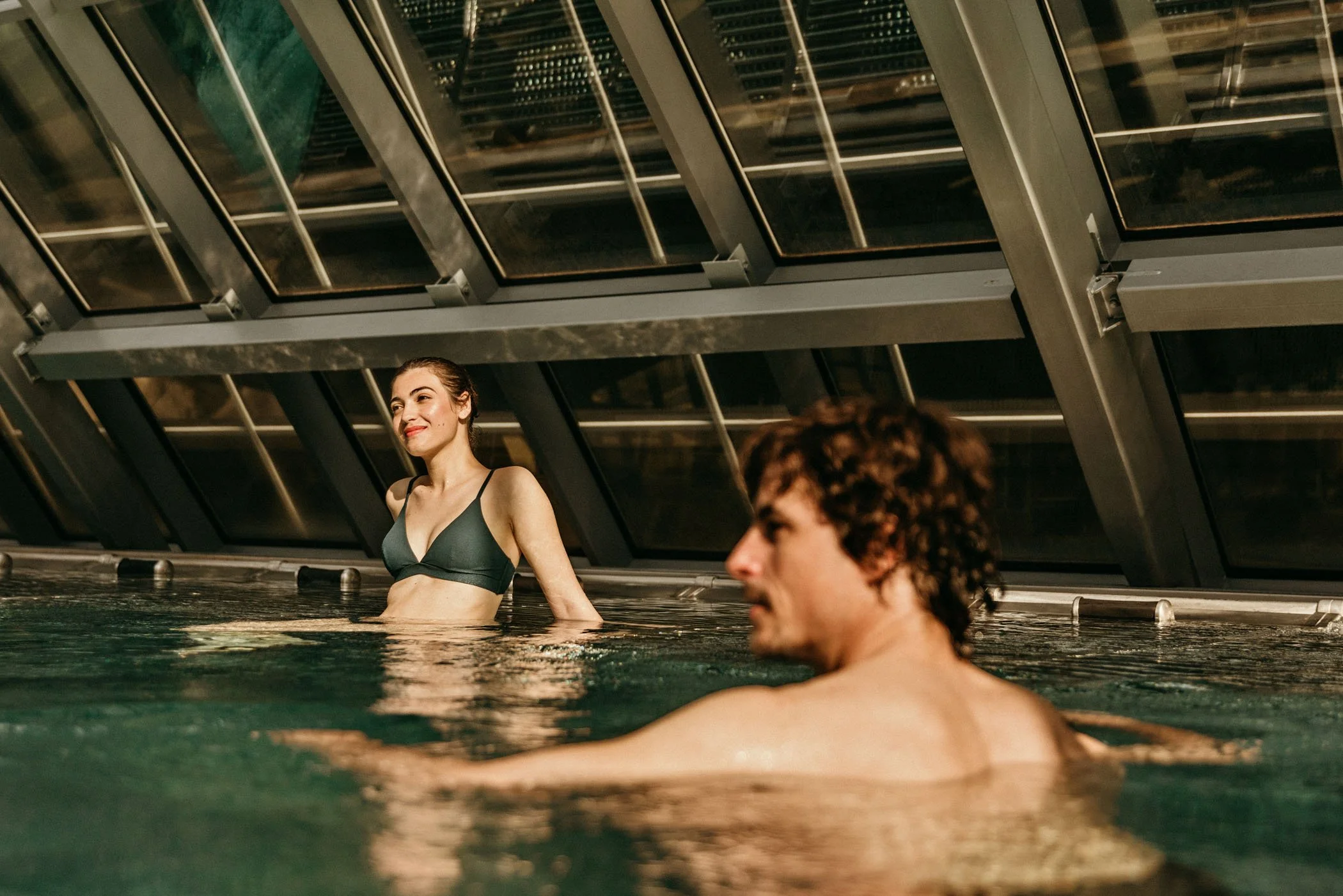 Una mujer sonriendo en la piscina, con un hombre en primer plano, en un entorno moderno y techado de cristales.