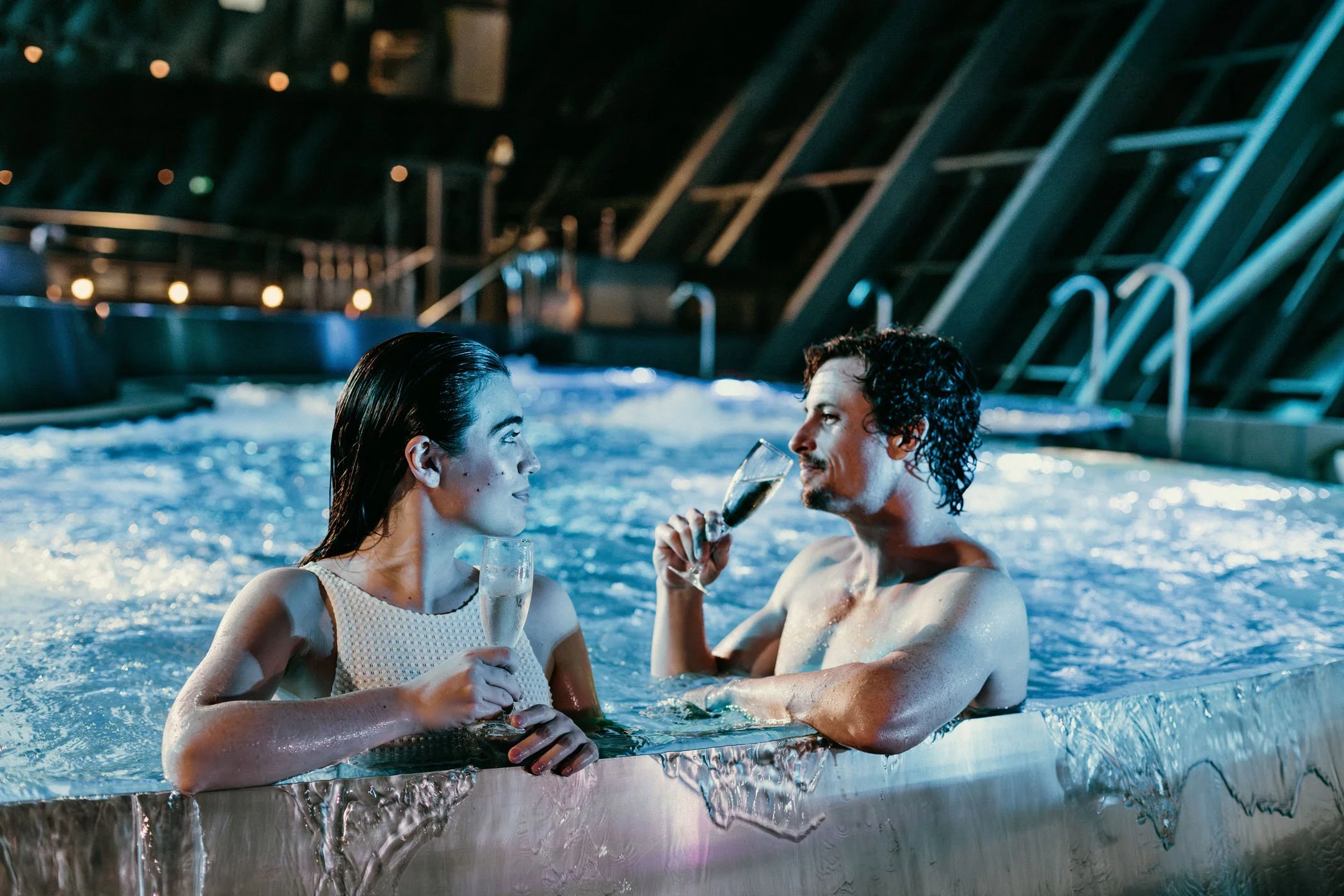 Pareja en una bañera de hidromasaje en un spa, brindando con copas de vino y viendo a su alrededor, ambiente acogedor y moderno con iluminación suave.