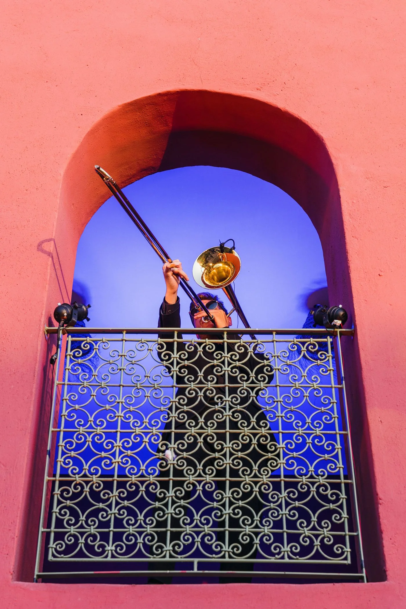 Músico tocando trombón desde un balcón con barandilla decorativa en un edificio rosa, con fondo azul y iluminación teatral.