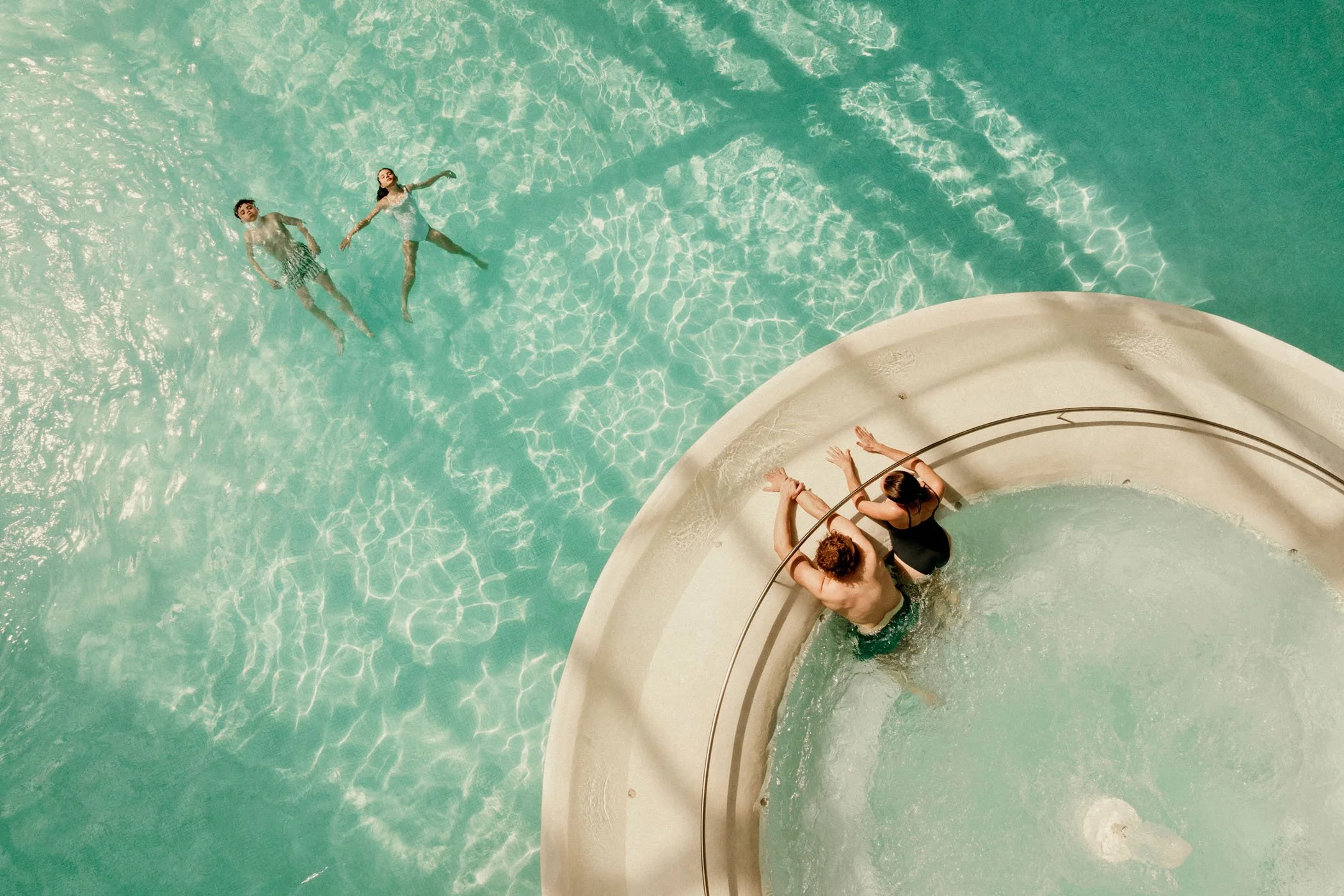 Dos personas en la alberca, una mujer y un hombre, y dos niños flotando en el agua, vista desde arriba.
