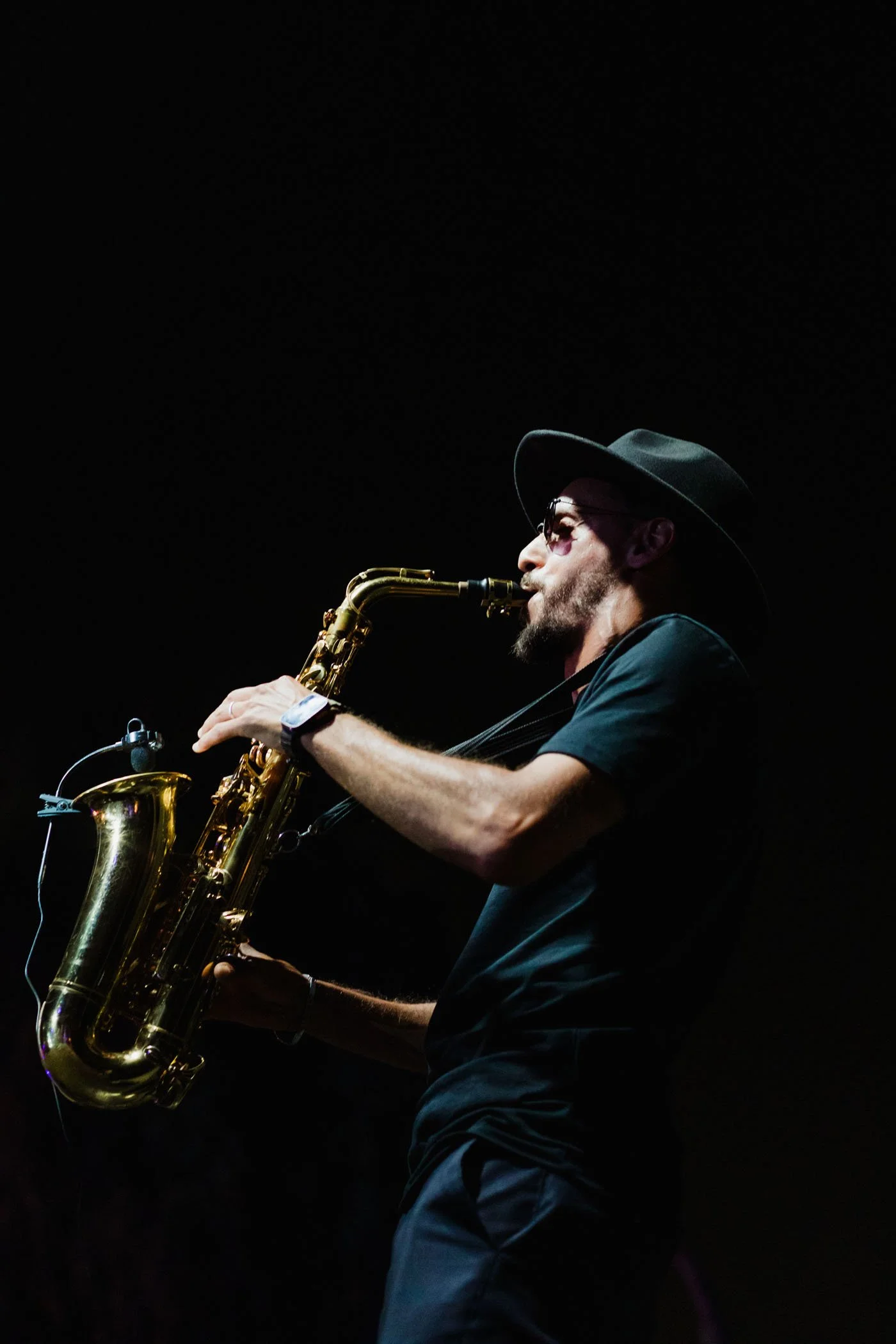 Hombre tocando el saxofón en un escenario oscuro, lleva sombrero y gafas de sol.