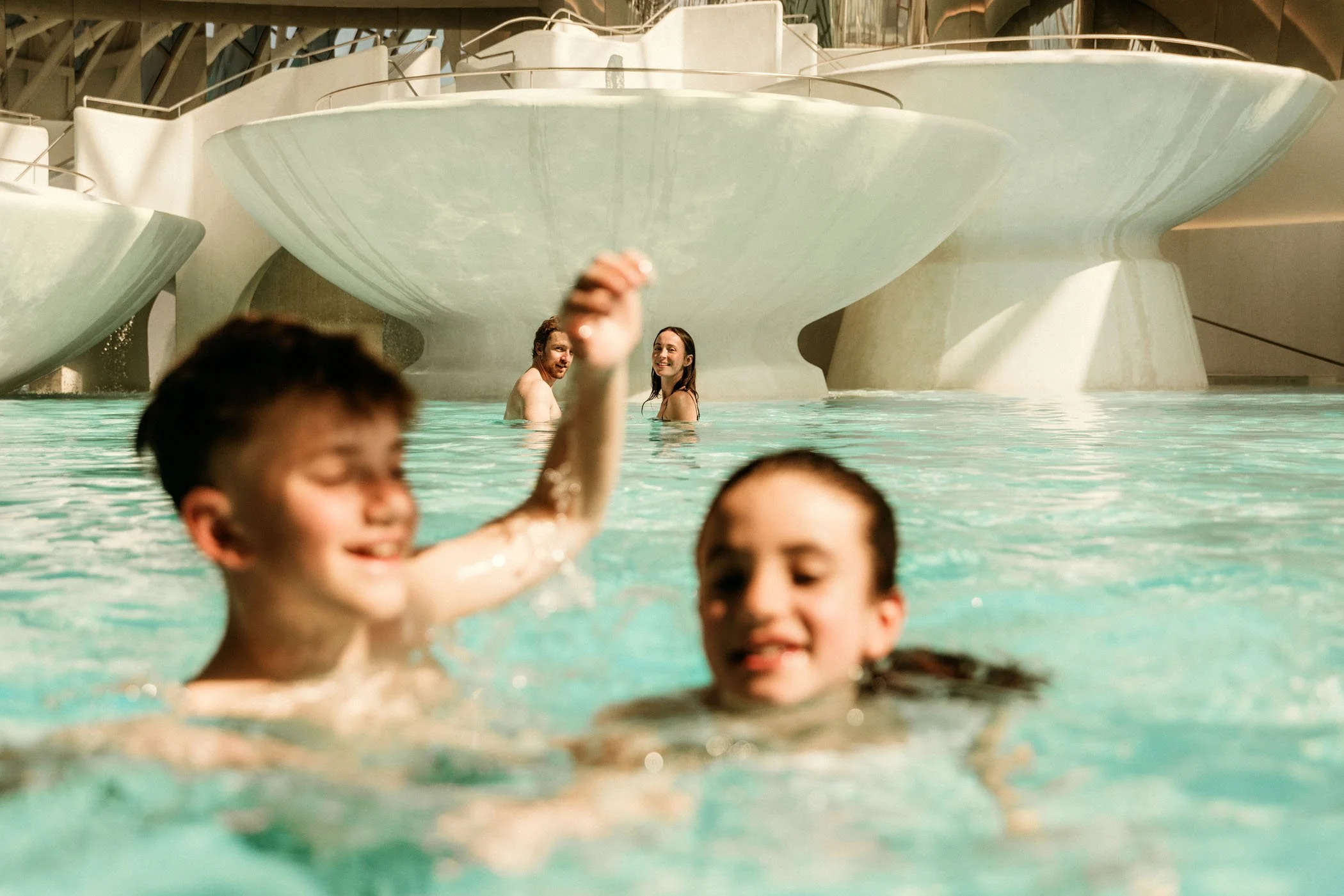 Niños jugando en una alberca con adultos disfrutando en el fondo, en un parque acuático con toboganes grandes y estructuras de agua.