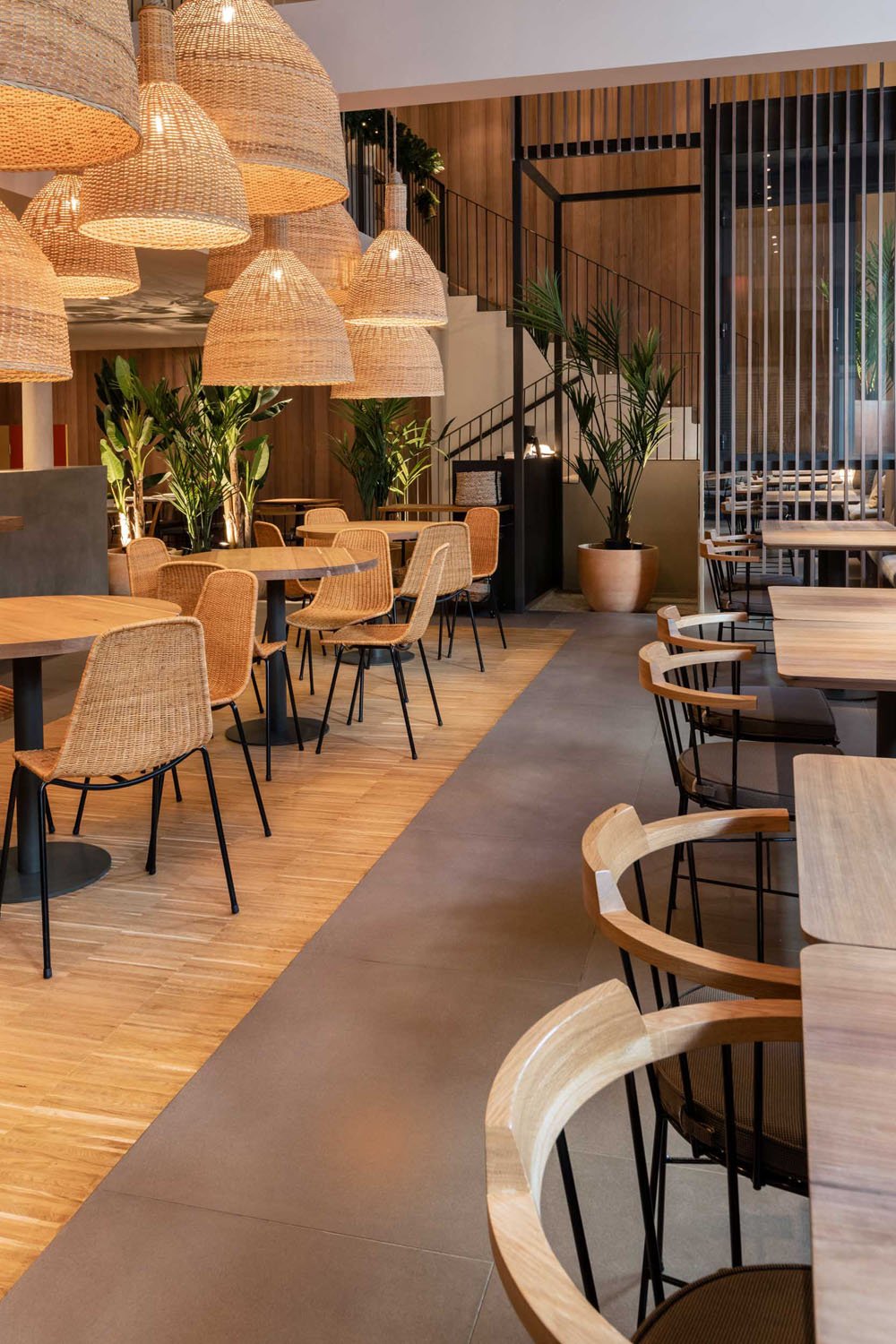 Interior de un restaurante con sillas y mesas de madera, lámparas de mimbre colgantes, plantas verdes en macetas grandes, paredes de madera y una escalera metálica en el fondo.