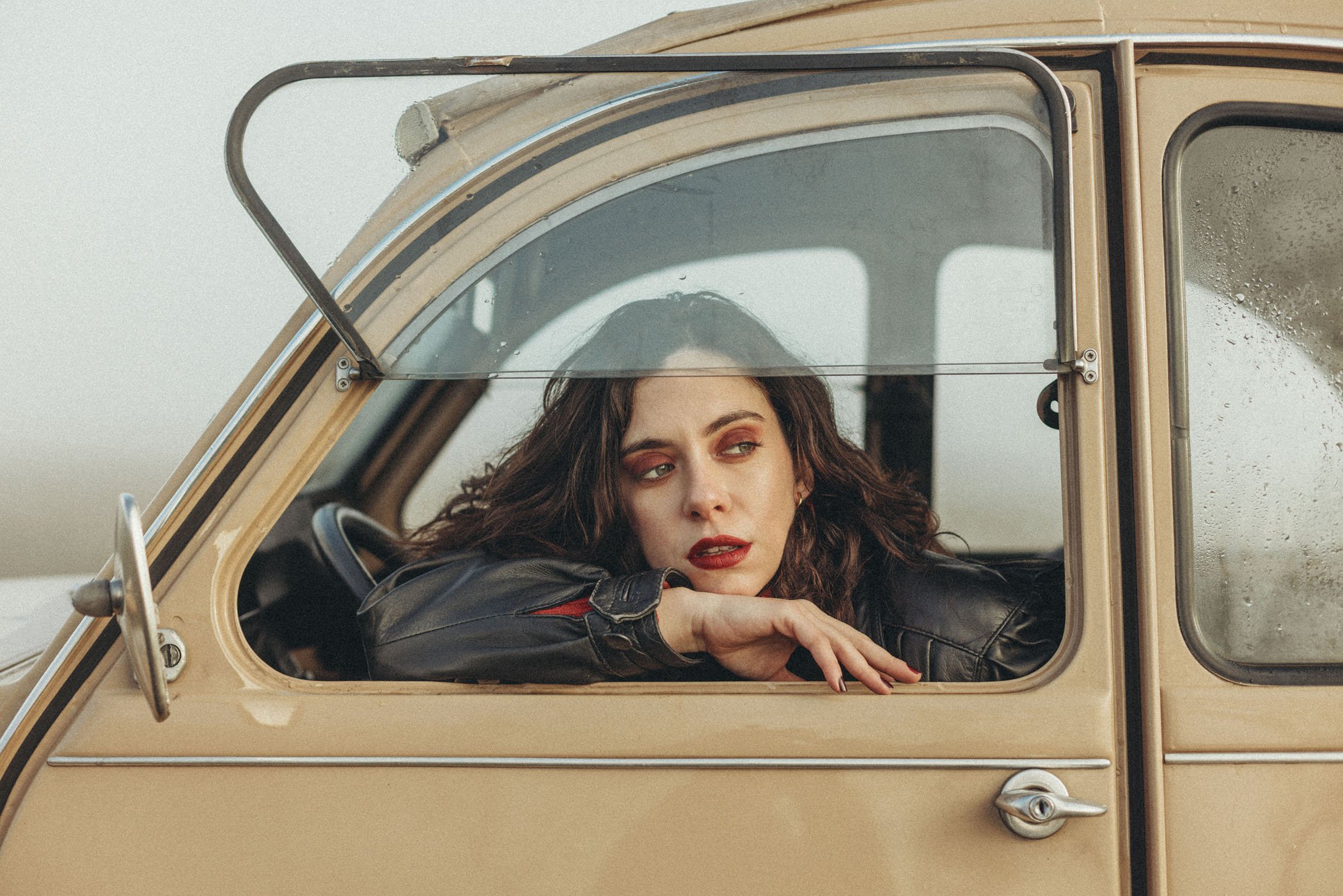 Mujer con cabello rizado y labios rojos mirando por la ventana de un coche clásico beige.