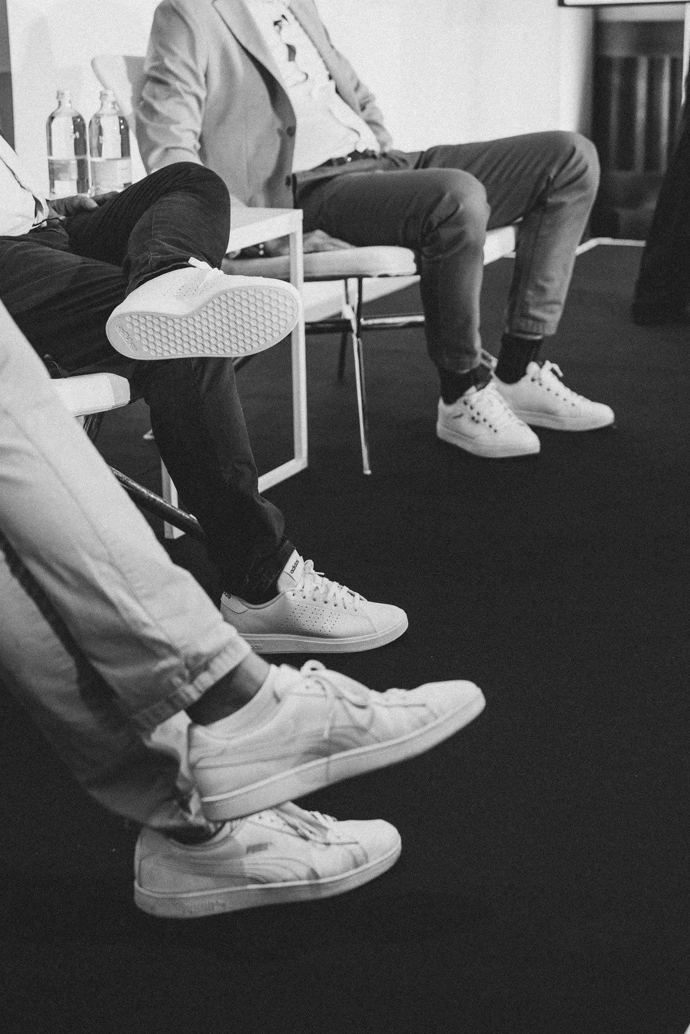 Imagen en blanco y negro de varias personas sentadas, enfocado en sus piernas y zapatos deportivos blancos.
