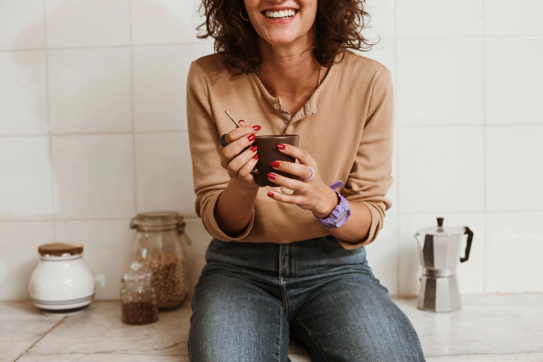 Mujer sonriendo mientras sostiene una taza en una cocina moderna, con frascos de condimentos y una cafetera en el fondo.