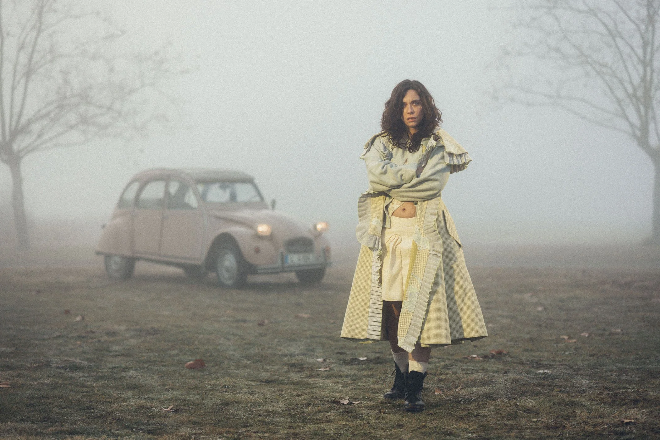 Mujer con ropa beige y grisada en un campo con niebla, con un coche antiguo y árboles desnudos en el fondo.
