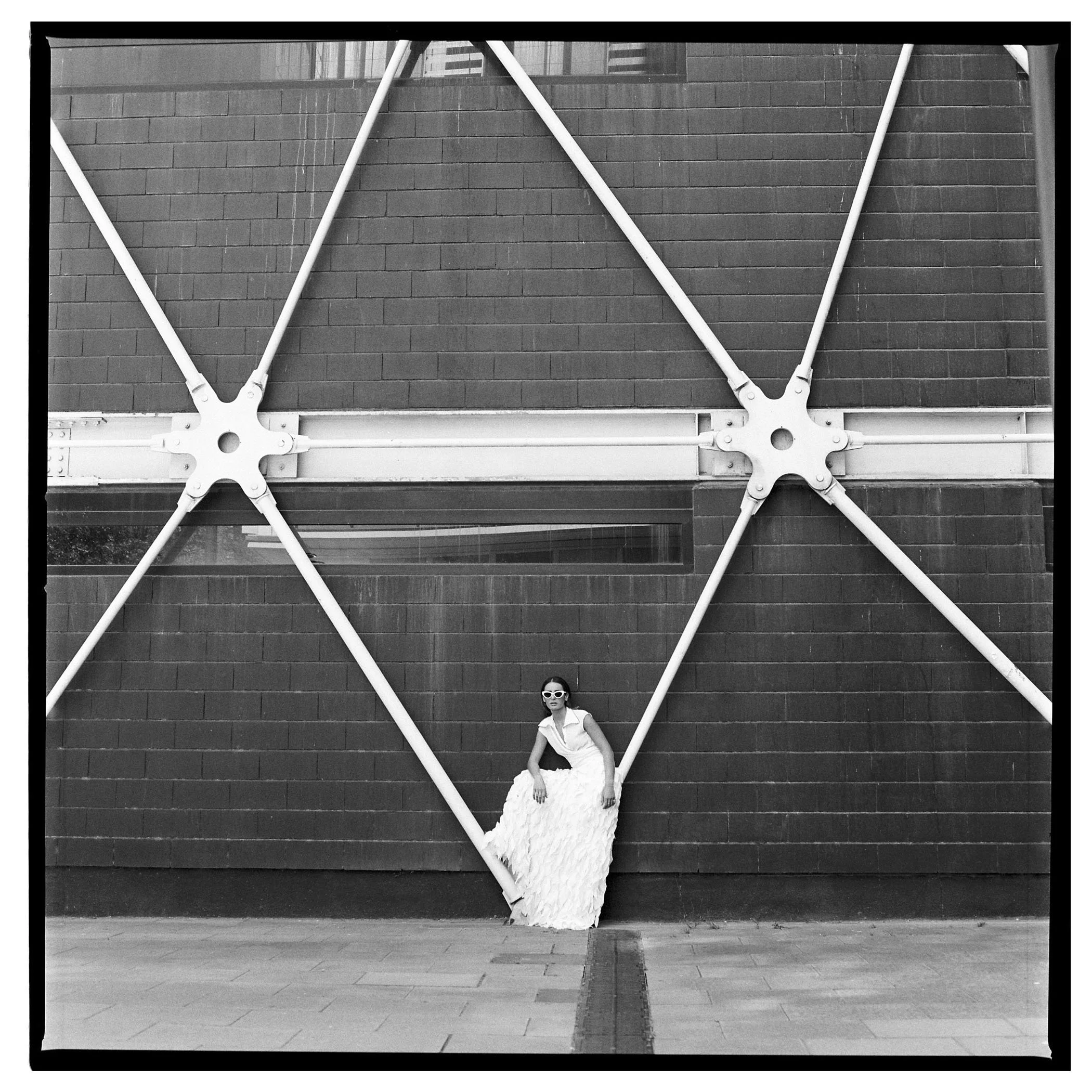Una mujer con vestido largo y gafas blancas sentada frente a un muro con estructura metálica en un entorno urbano.