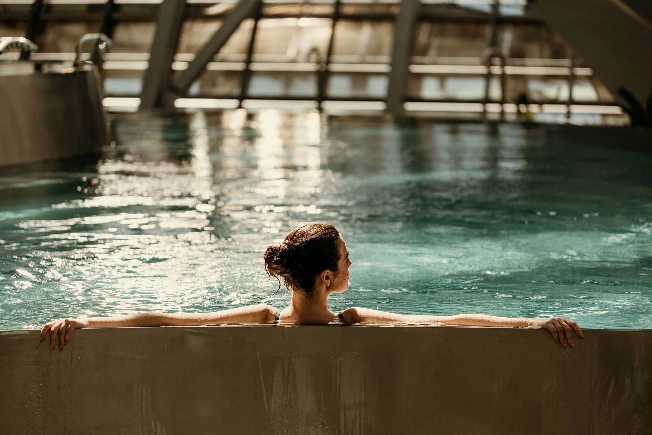 Mujer en una piscina cubierta, descansando con los brazos extendidos por el borde y mirando hacia un lado, con estructura metálica y ventanas en el fondo.