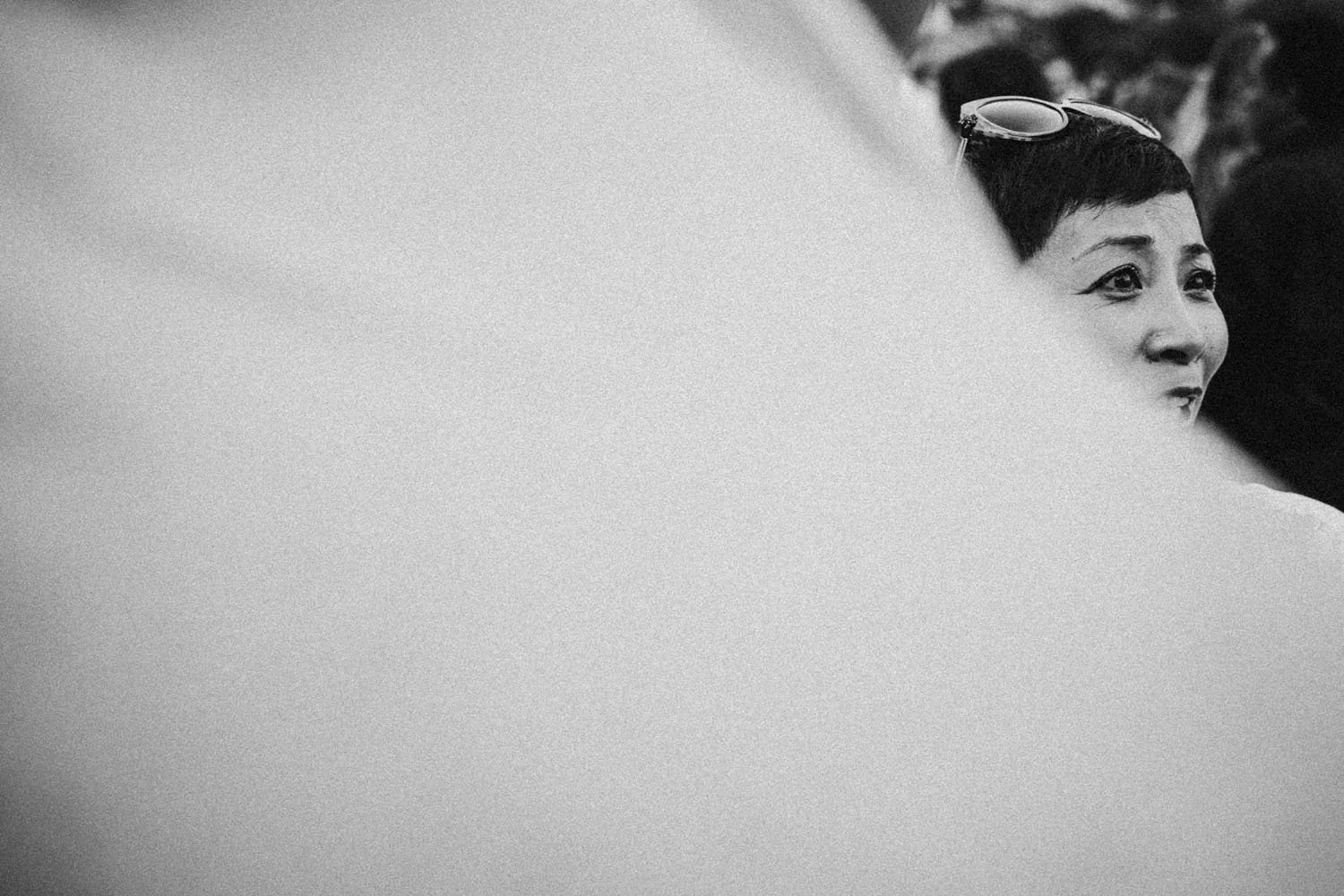 Retrato en blanco y negro de una mujer con gafas en la cabeza, mirando hacia la derecha y sonriendo ligeramente, parcialmente oculta por una superficie borrosa en primer plano.