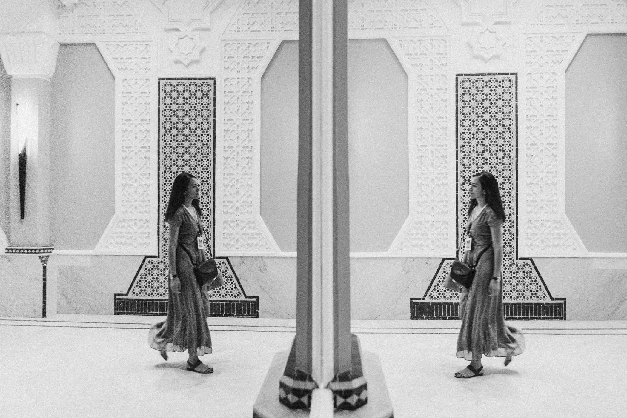 Una mujer con vestido largo y bolso camina por un pasillo adornado con patrones geométricos en las paredes, vista reflejada en un espejo central.