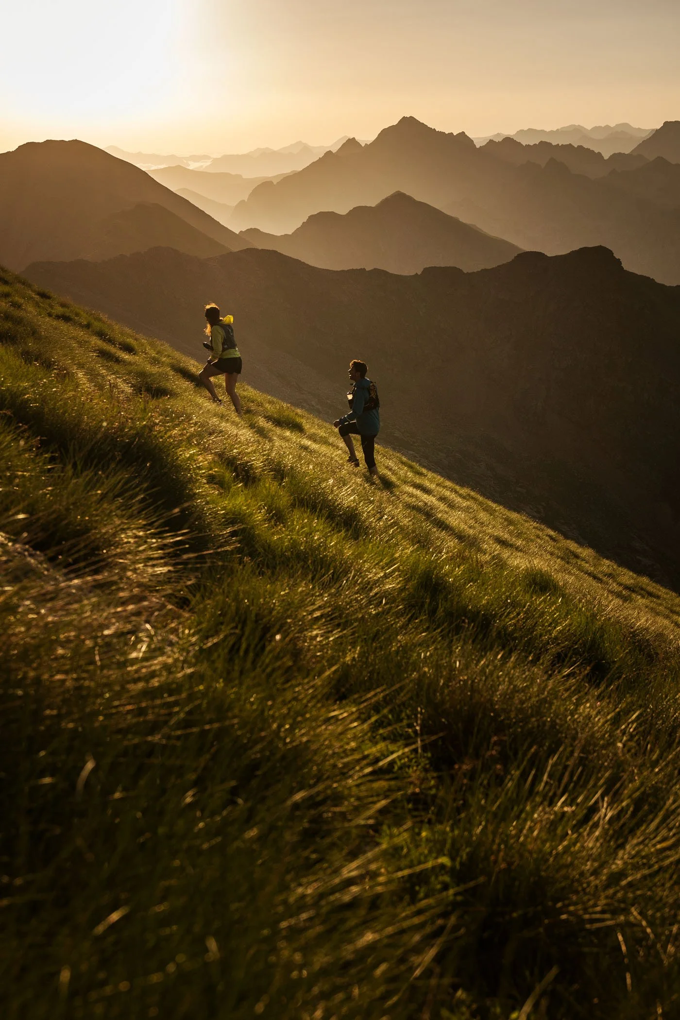 Dos personas haciendo senderismo en una ladera de montaña durante la puesta del sol, con un paisaje de montañas al fondo.