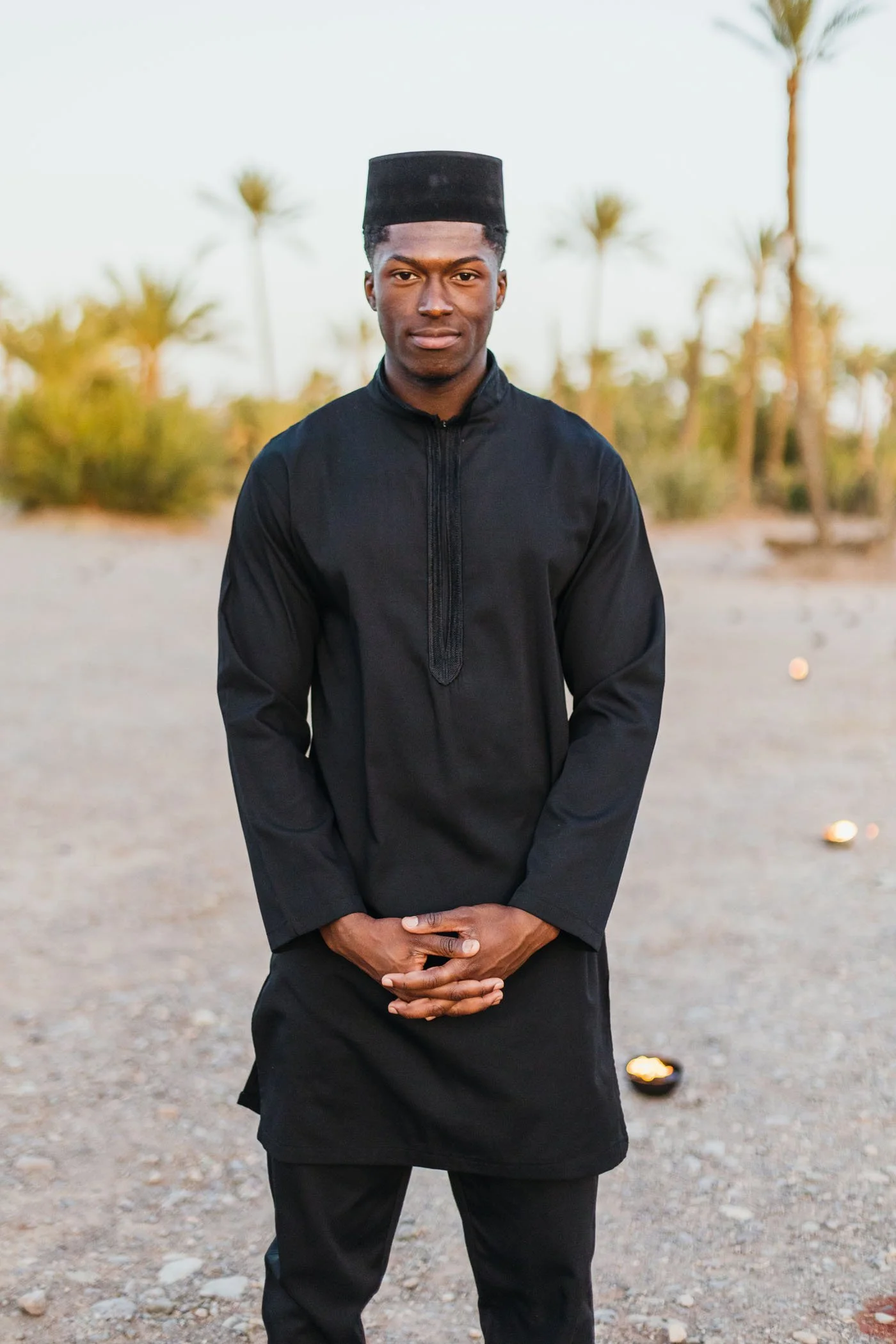 Joven hombre de pie en un entorno desértico con palmeras, vestido con ropa tradicional negra y tocado negro.