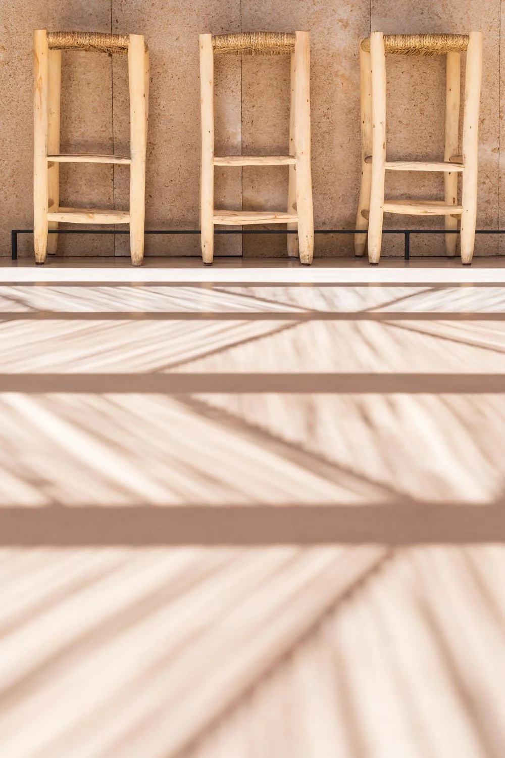 Tres sillas de madera de diseño rústico frente a una pared de piedra, con sombra de hojas en el suelo.