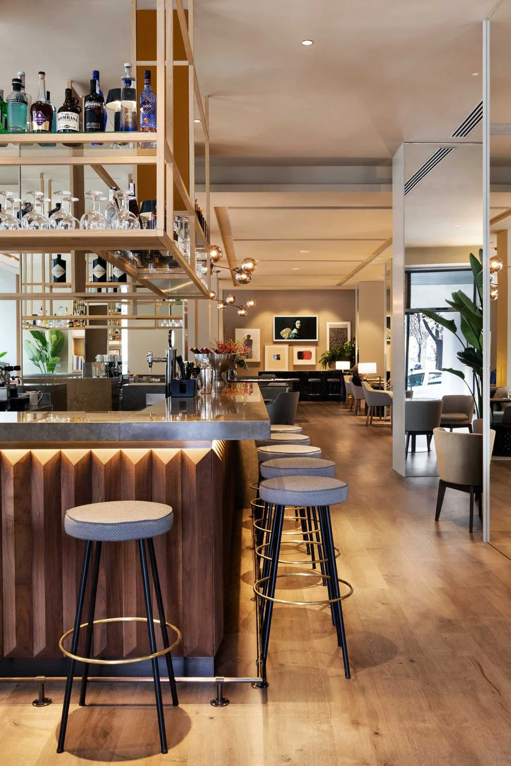 interior de un restaurante elegante con barra de madera, taburetes de respaldo bajo con asientos de tela gris, decoraciones modernas y plantas verdes, iluminación cálida y paredes decoradas con cuadros