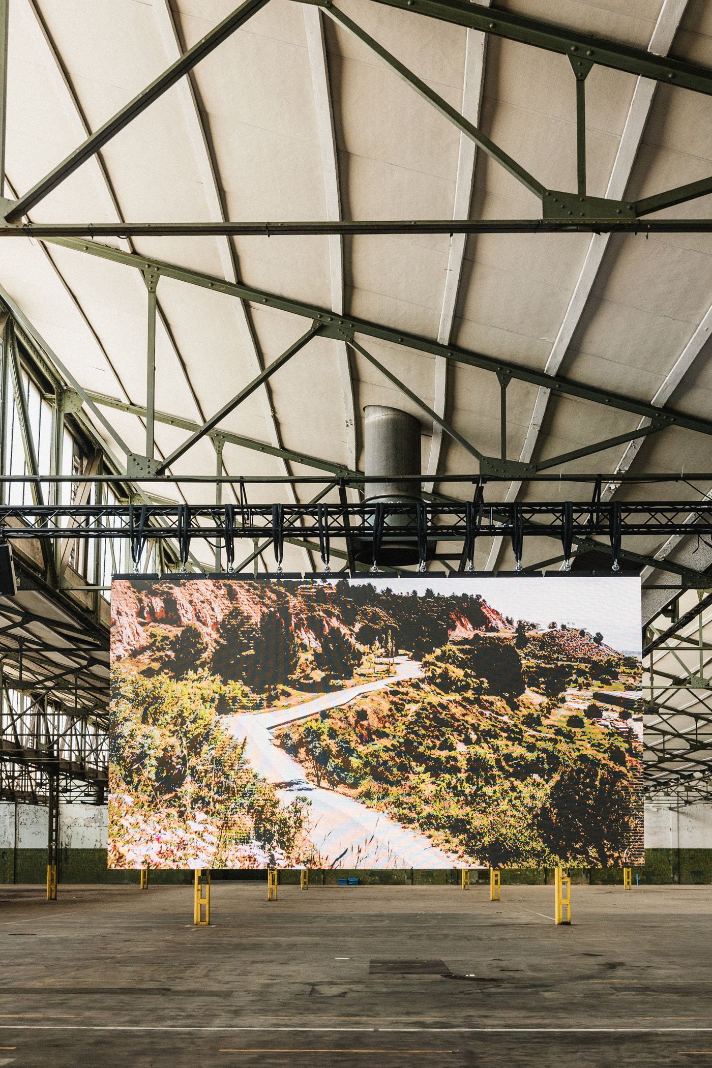 Una pantalla gigante en el interior de un espacio industrial que muestra una vista panorámica de una carretera que atraviesa un paisaje montañoso con vegetación.