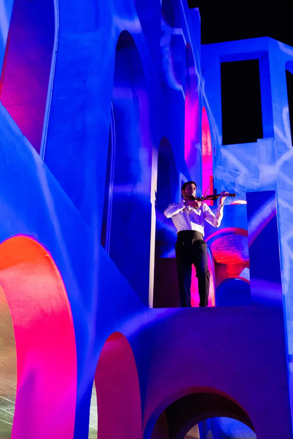 Hombre tocando violín en escenario con decoraciones azules, rosas y púrpuras en forma de arcos y estructuras arquitectónicas.