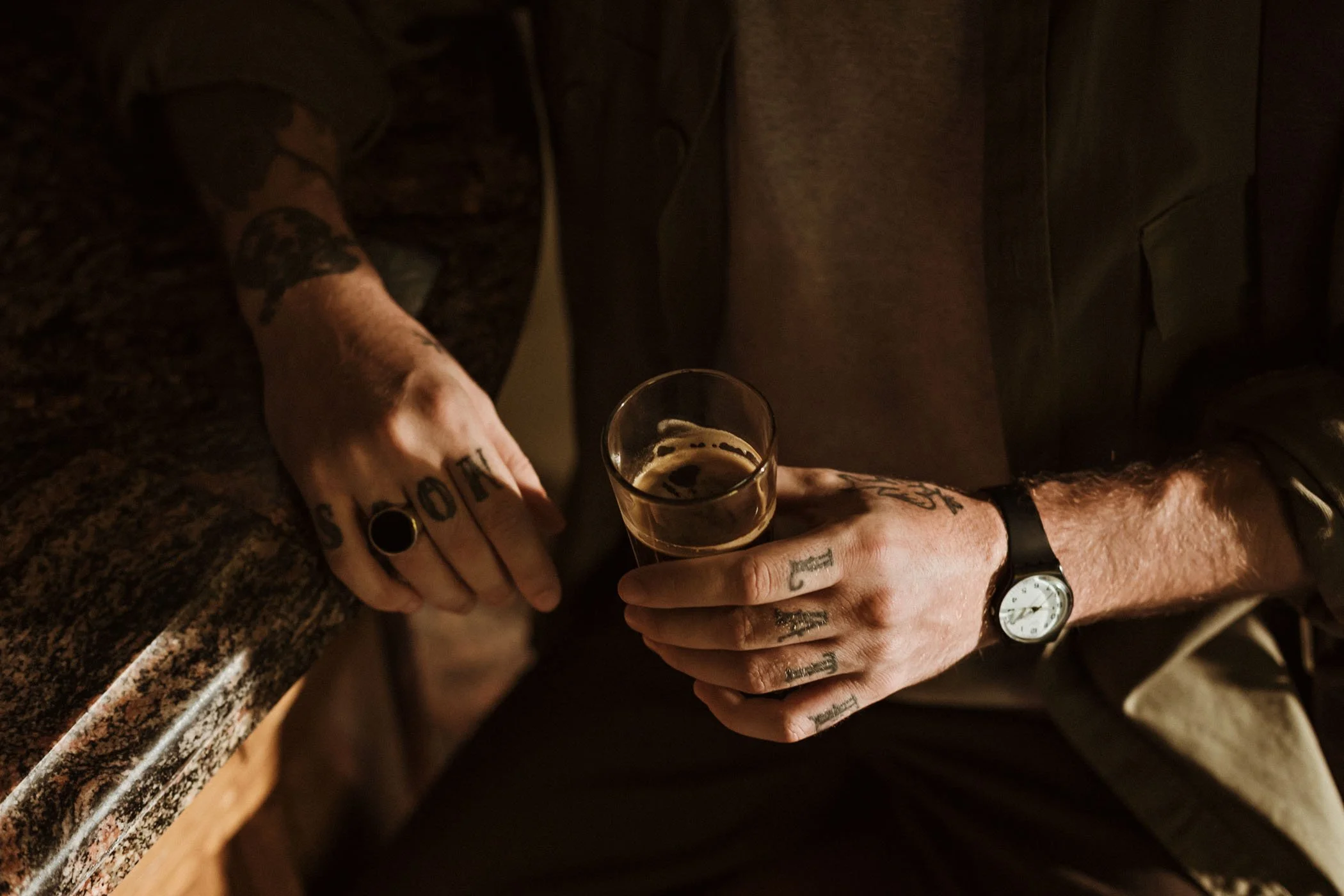 Persona con tatuajes sosteniendo un vaso de cerveza en una mano, lleva un reloj y un anillo en el dedo, y está apoyada en una barra de granito en un ambiente oscuro.