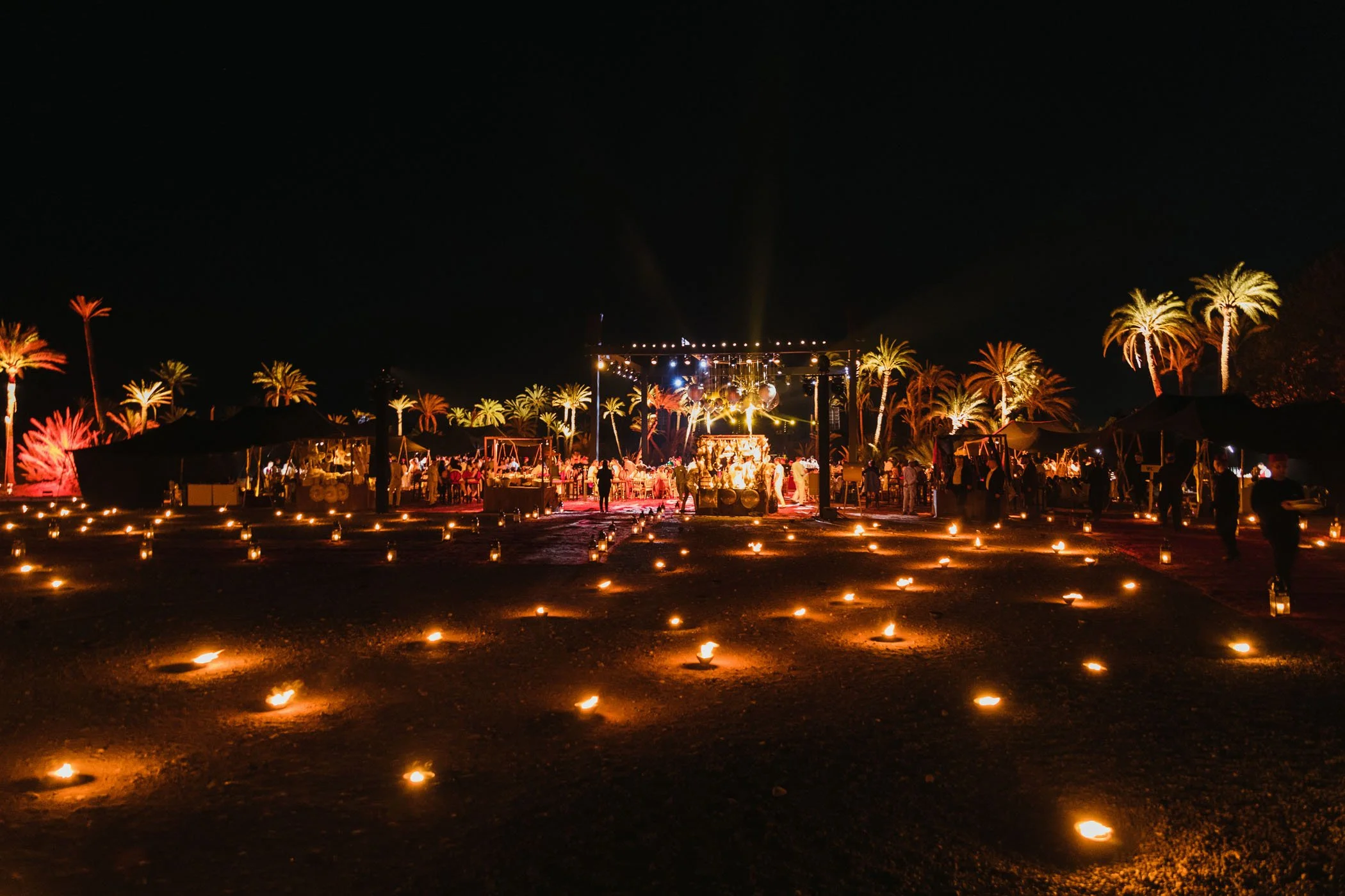 Una celebración nocturna con varias velas encendidas en el suelo, un escenario iluminado con música, muchas palmeras al fondo y varias personas disfrutando del evento.