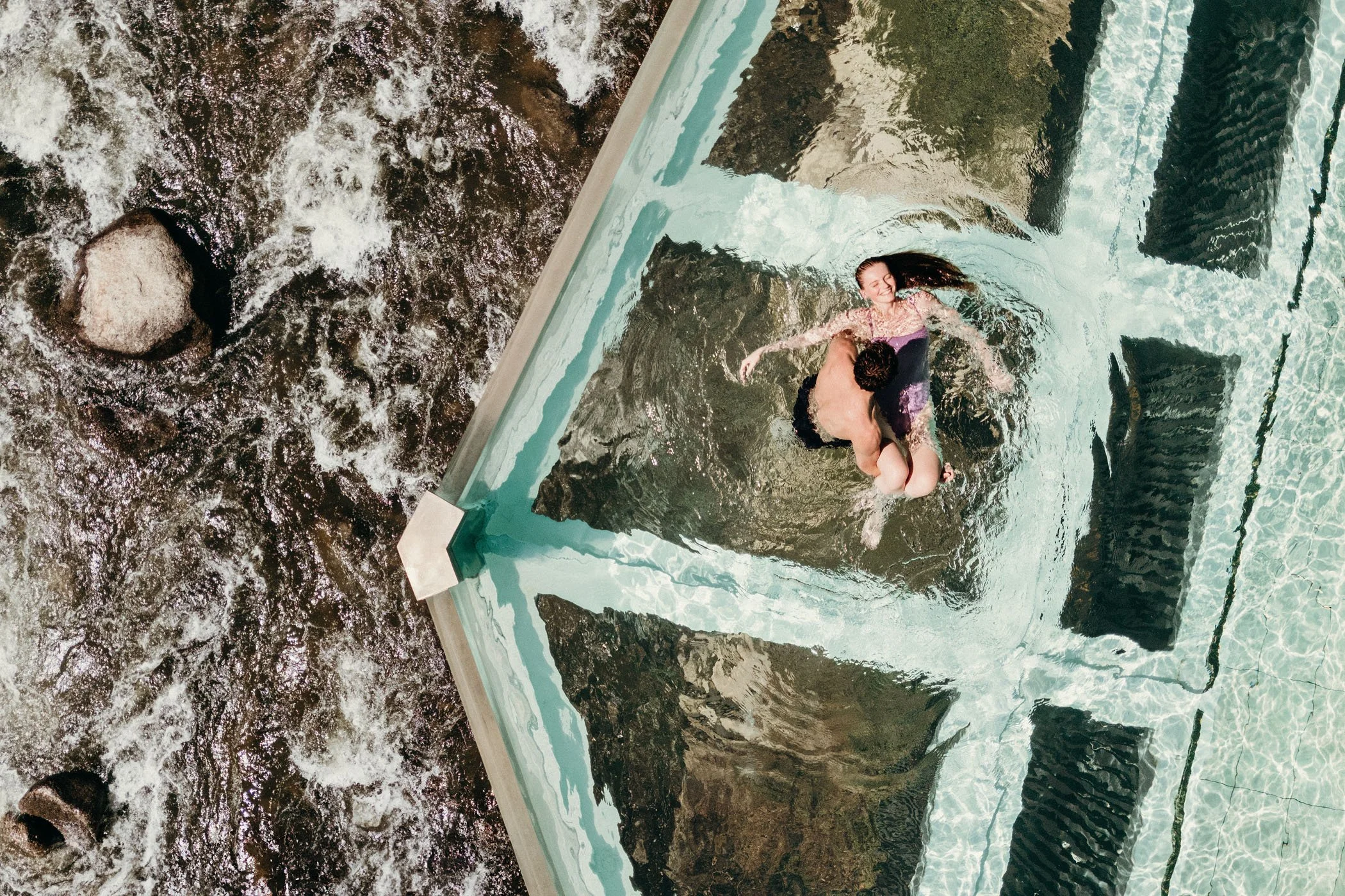 Pareja en una piscina con estructura de vidrio, riendo y abrazándose mientras la corriente del río fluye cerca.