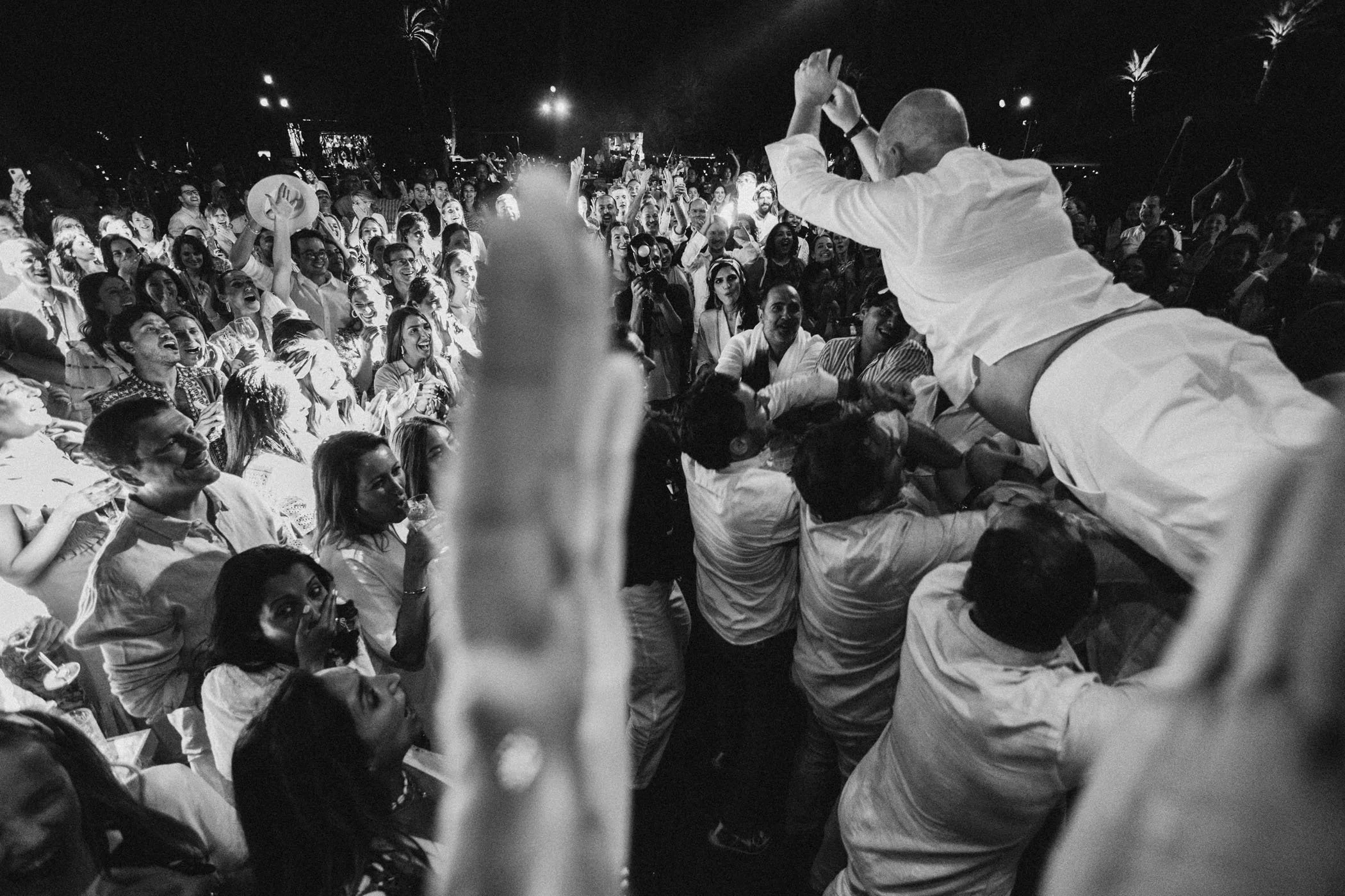 Hombre levantado en un dancón, rodeado de muchas personas alegres en un evento nocturno al aire libre.