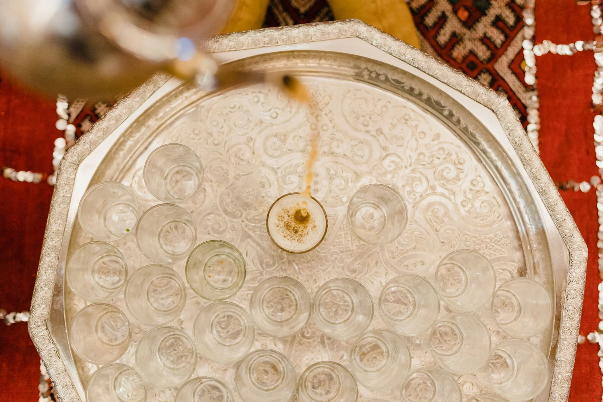 Vista desde arriba de una bandeja con varios vasos de cristal y una fuente de bebida en el centro, sobre un tapete decorativo rojo y negro.