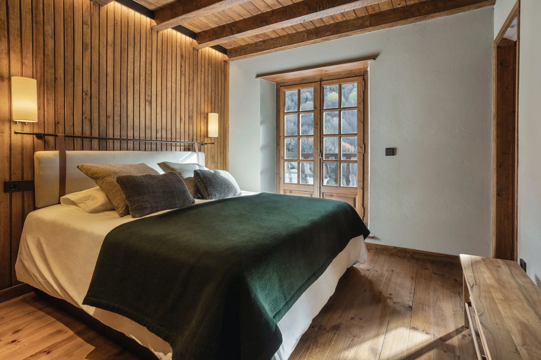 Recámara con paredes de madera, ventanas de madera con vista a la naturaleza, cama con cobija verde y cojines grises, lámparas de pared