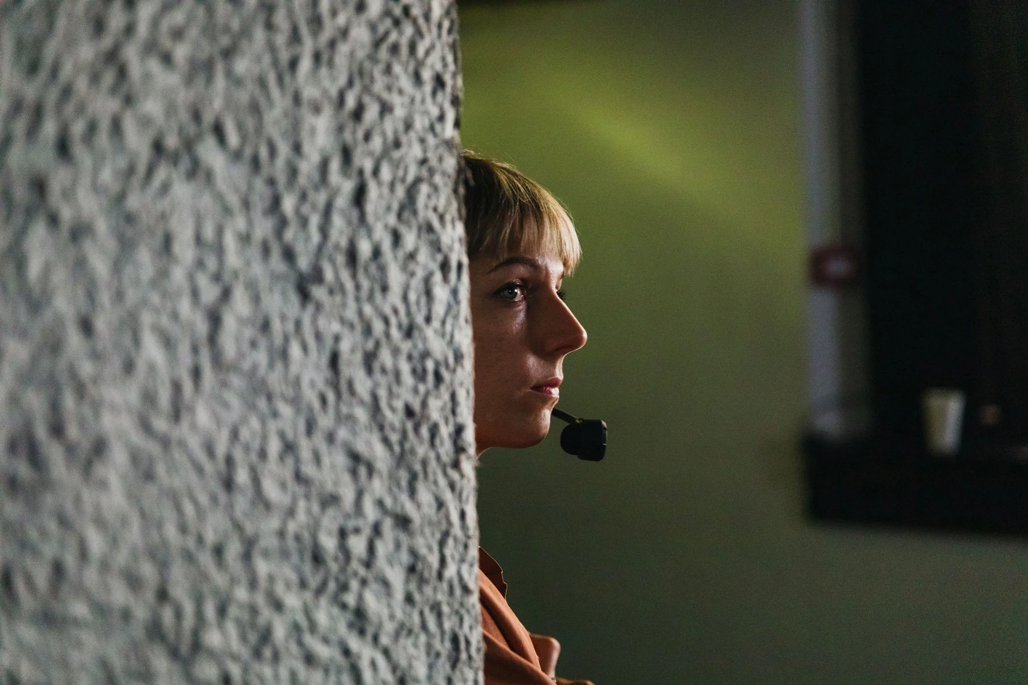 Mujer con auricular de micrófono viendo hacia la derecha, parcialmente oculta tras una pared de textura rugosa 