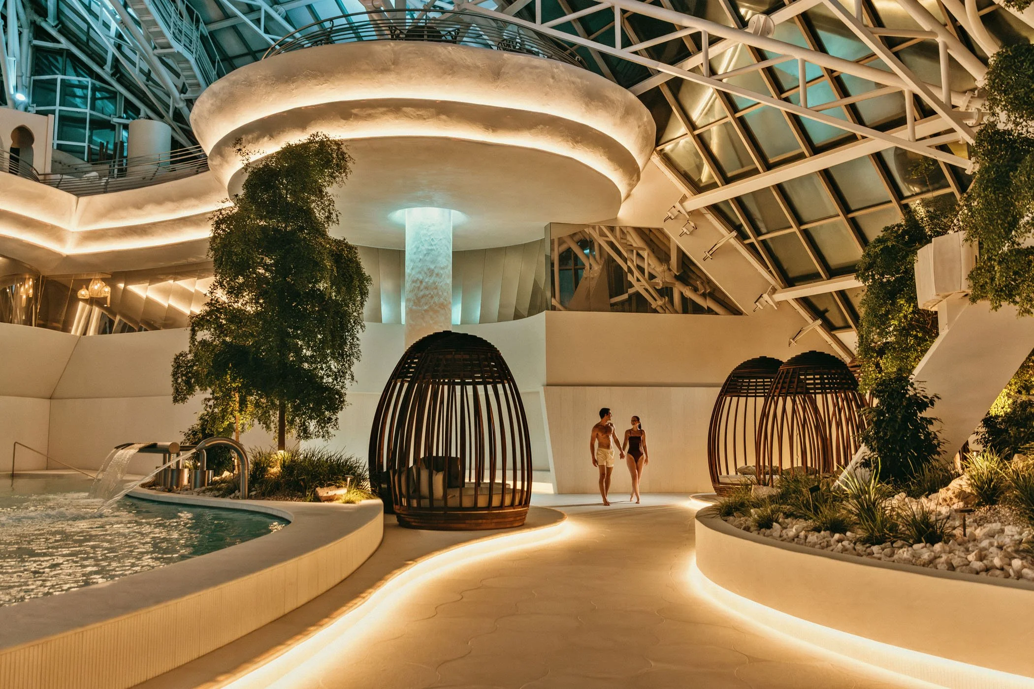 Interior moderno con zonas de descanso, árboles y muebles de cabaña en un espacio con iluminación cálida y estructura de vidrio y metal.