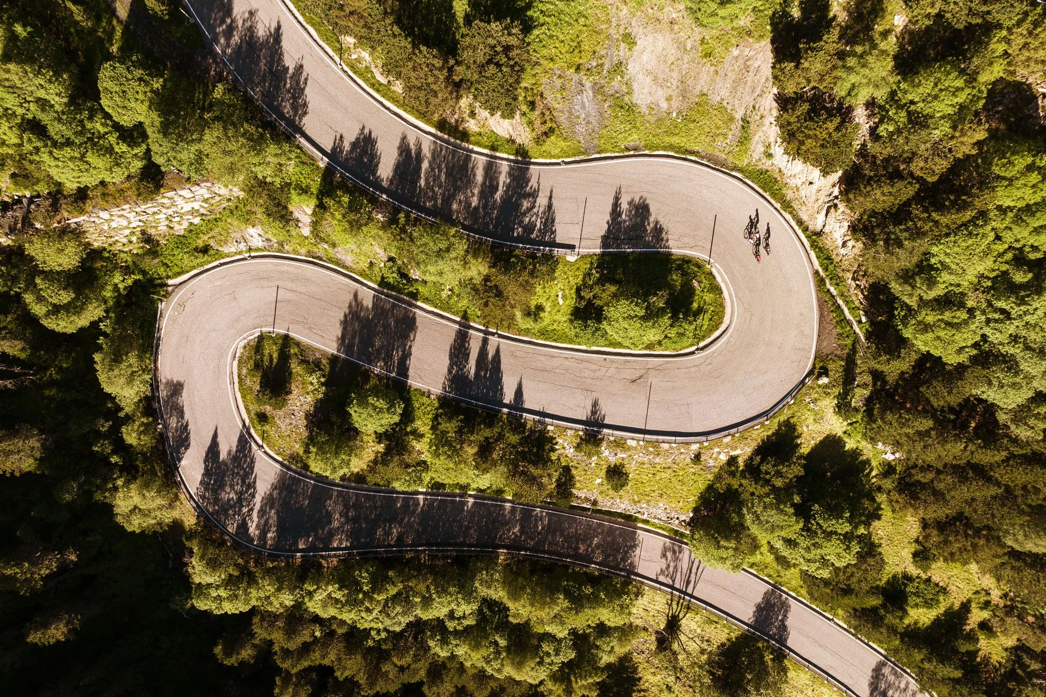 Vista aérea de una carretera en zigzag rodeada de árboles y vegetación, con dos ciclistas.