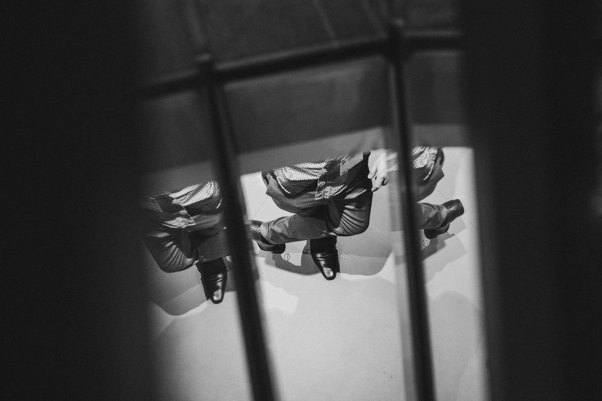 Vista en ángulo de una persona vestida con traje de negocios y zapatos oscuros, tomada desde arriba y a través de una estructura de barras, con sombra en el suelo.