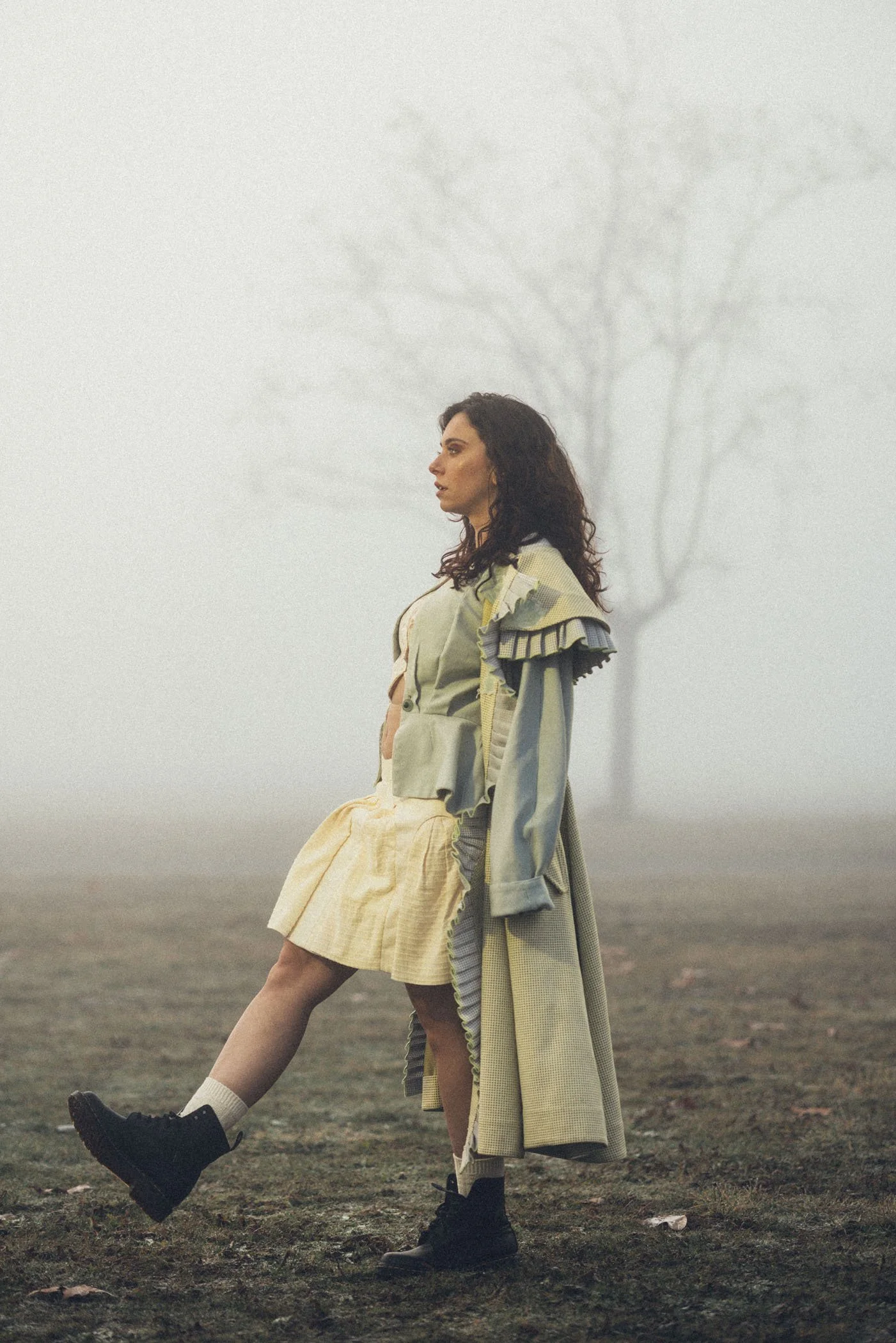 Mujer con vestido y abrigo largo en un campo con neblina y árbol sin hojas al fondo.
