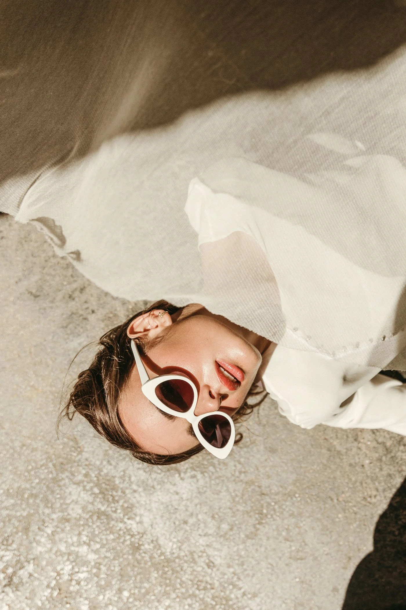 Mujer con gafas blancas y labios rojos acostada en la arena, parcialmente cubierta por una tela blanca.