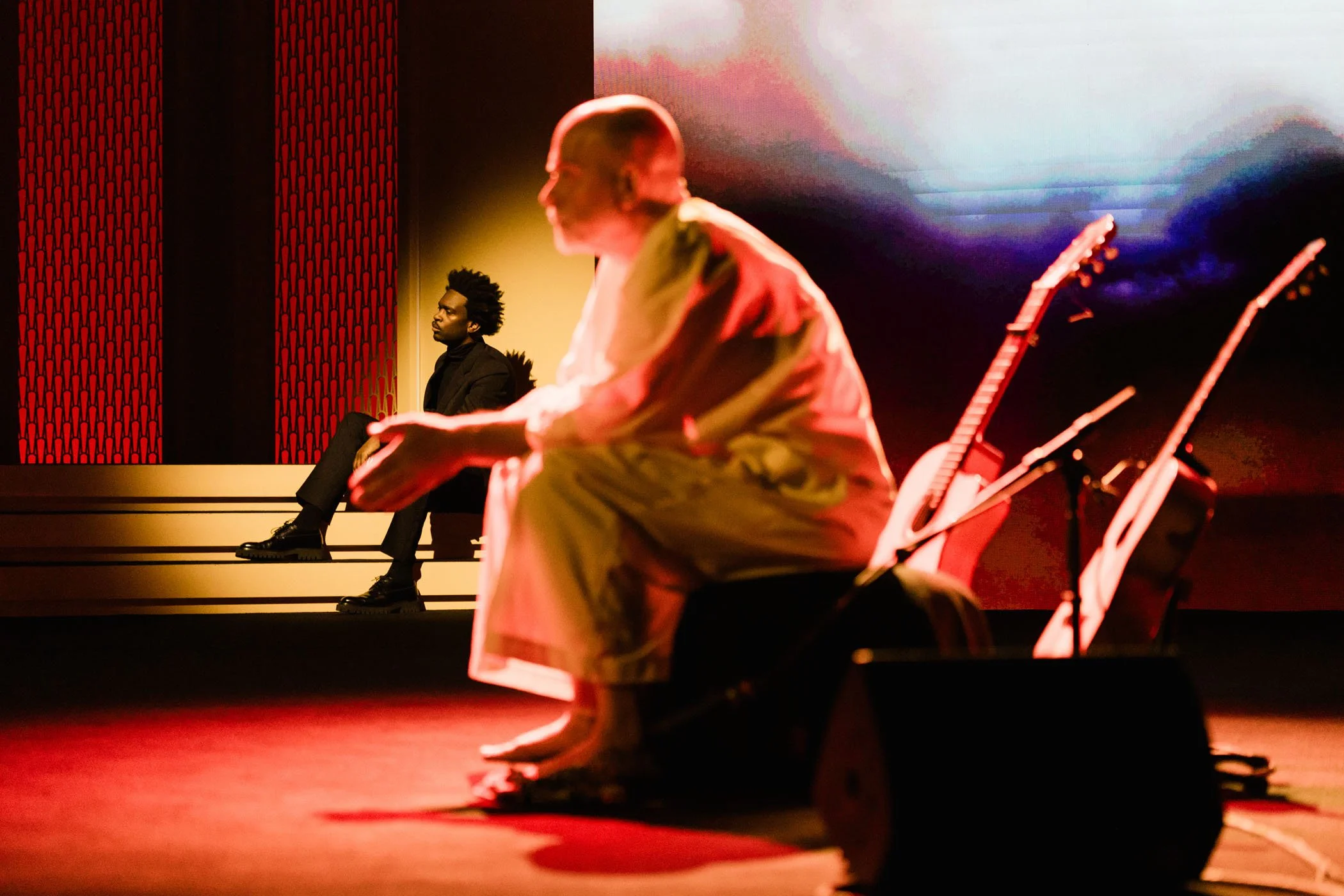Dos personas sentadas en un escenario con guitarras, uno en primer plano y otro en segundo plano, durante una presentación artística en un ambiente íntimo y con iluminación cálida.