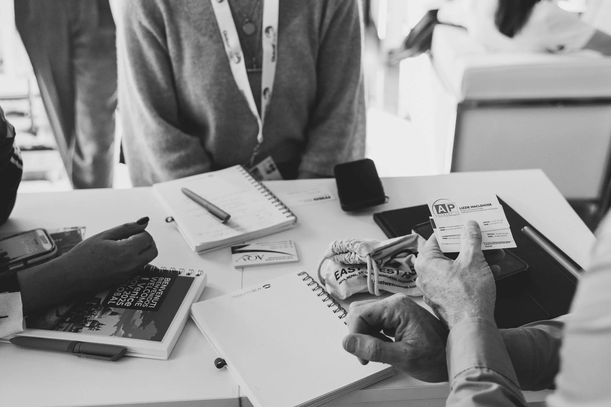 Personas intercambiando documentos y notas en una mesa de oficina, con objetos como libretas, una tarjeta de presentación, un teléfono móvil, y una bolsa con logotipo, en un ambiente de trabajo.