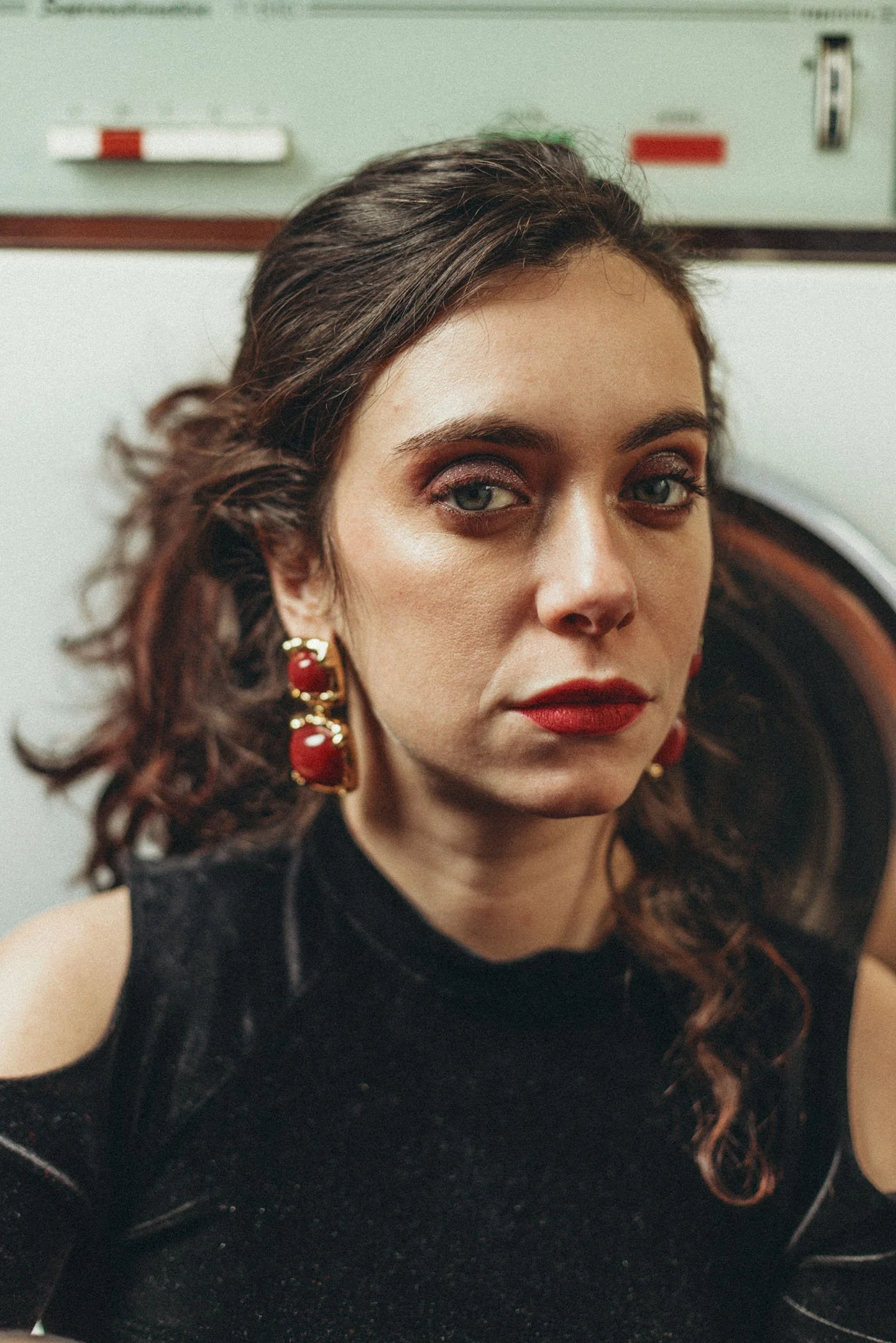 Retrato de una mujer con cabello rizado, maquillaje de ojos y labial rojo, usando aretes grandes con detalles dorados y rojos, con fondo que parece ser una lavandería o lavadora.
