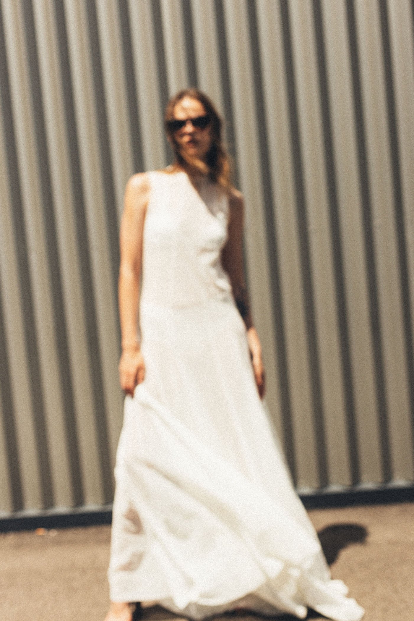 Mujer con vestido blanco largo y gafas de sol posando frente a una pared metálica vertical.