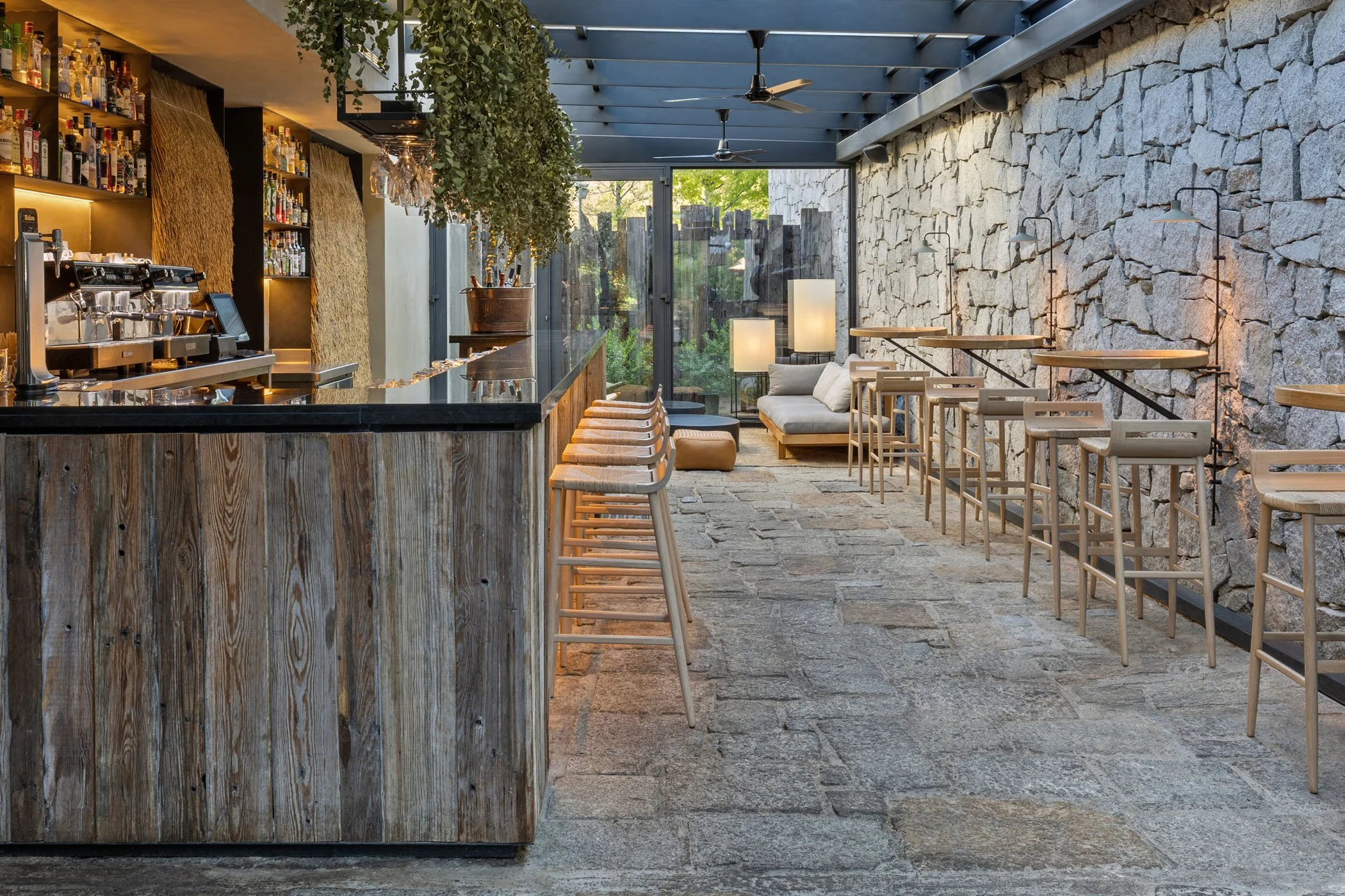 Interiores de un bar o cafetería con barra de madera, sillas de bar alineadas, una pared de piedra, iluminación cálida y áreas de descanso con sillones y lámparas, decorado con plantas y ventanales grandes que dejan ver vegetación afuera.