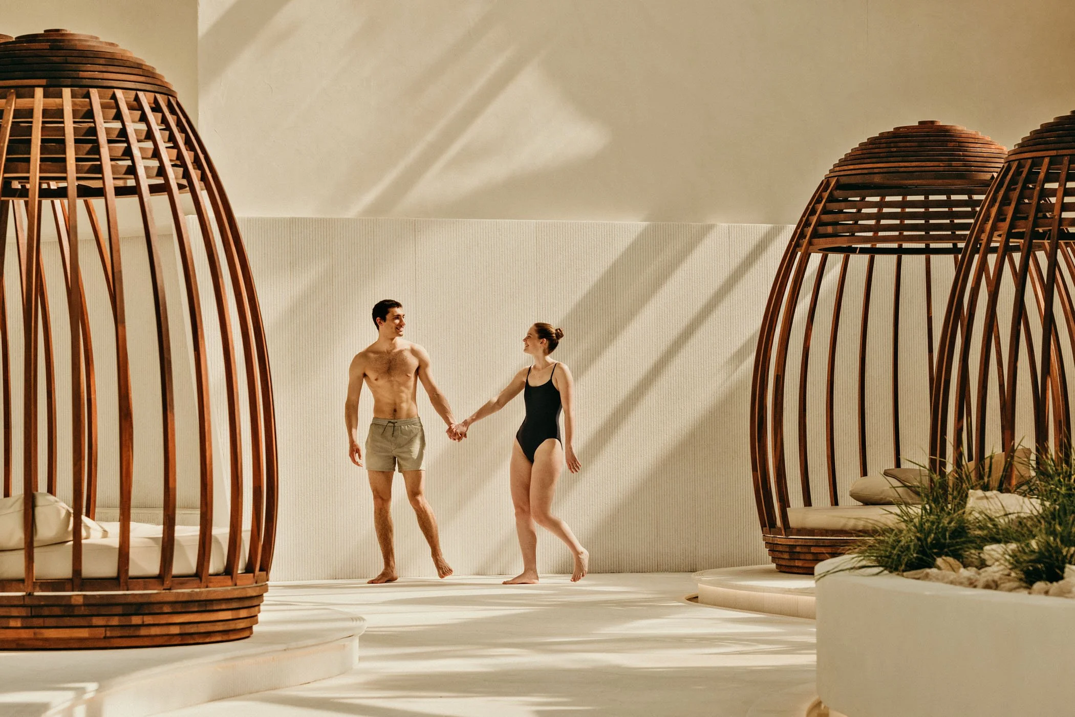 Una pareja joven de pie en una piscina, sujetándose de las manos, rodeados de cabañas de madera con sombra del sol en un ambiente moderno y relajante.