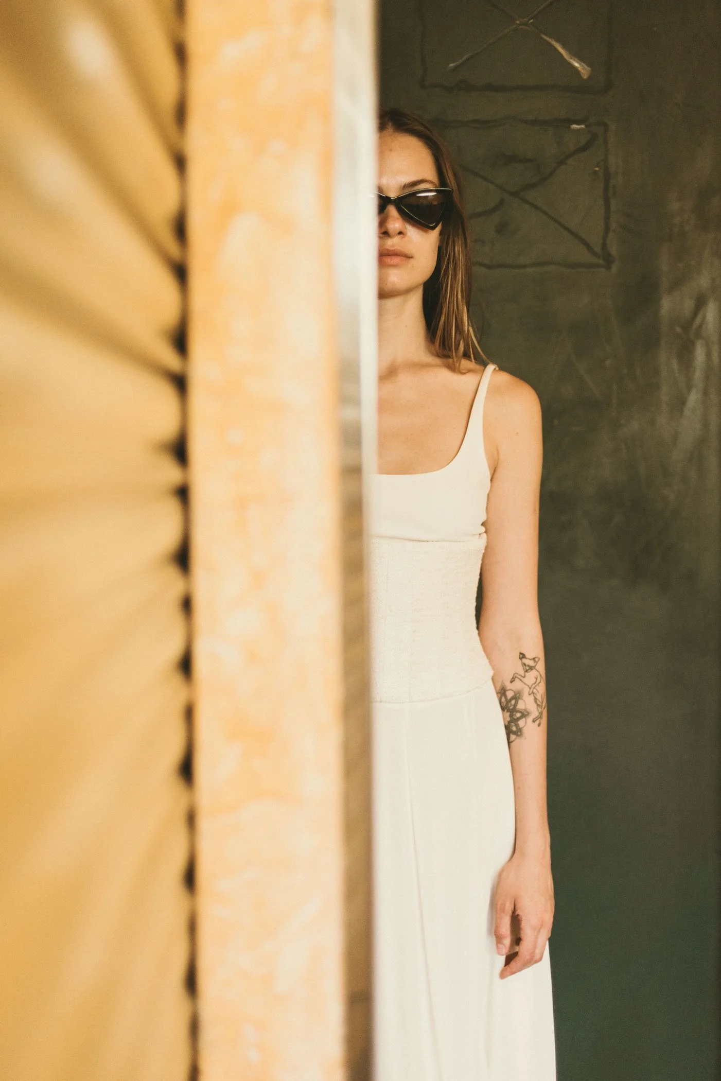 Mujer con gafas oscuras y vestido blanco, parcialmente visible, de pie junto a una pared gris y una estructura amarilla en un espacio decorado minimalista.