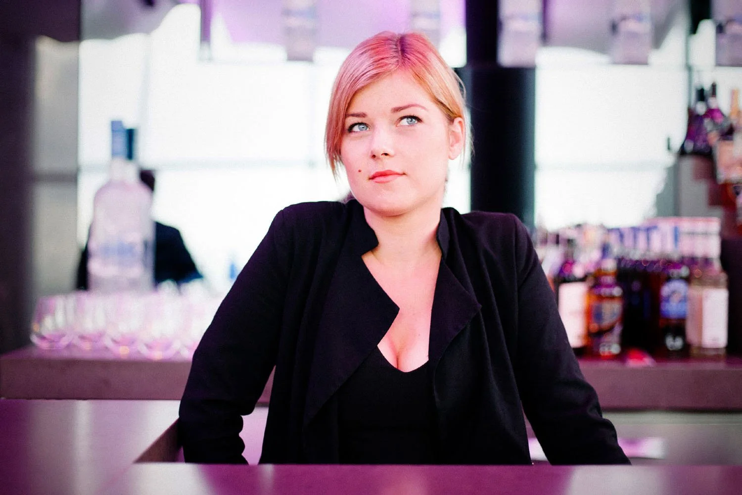 Una mujer con cabello rubio y de tez clara, vestida con ropa negra, sentada en una mesa en lo que parece un restaurante o cafetería, con una expresión pensativa o relajada.