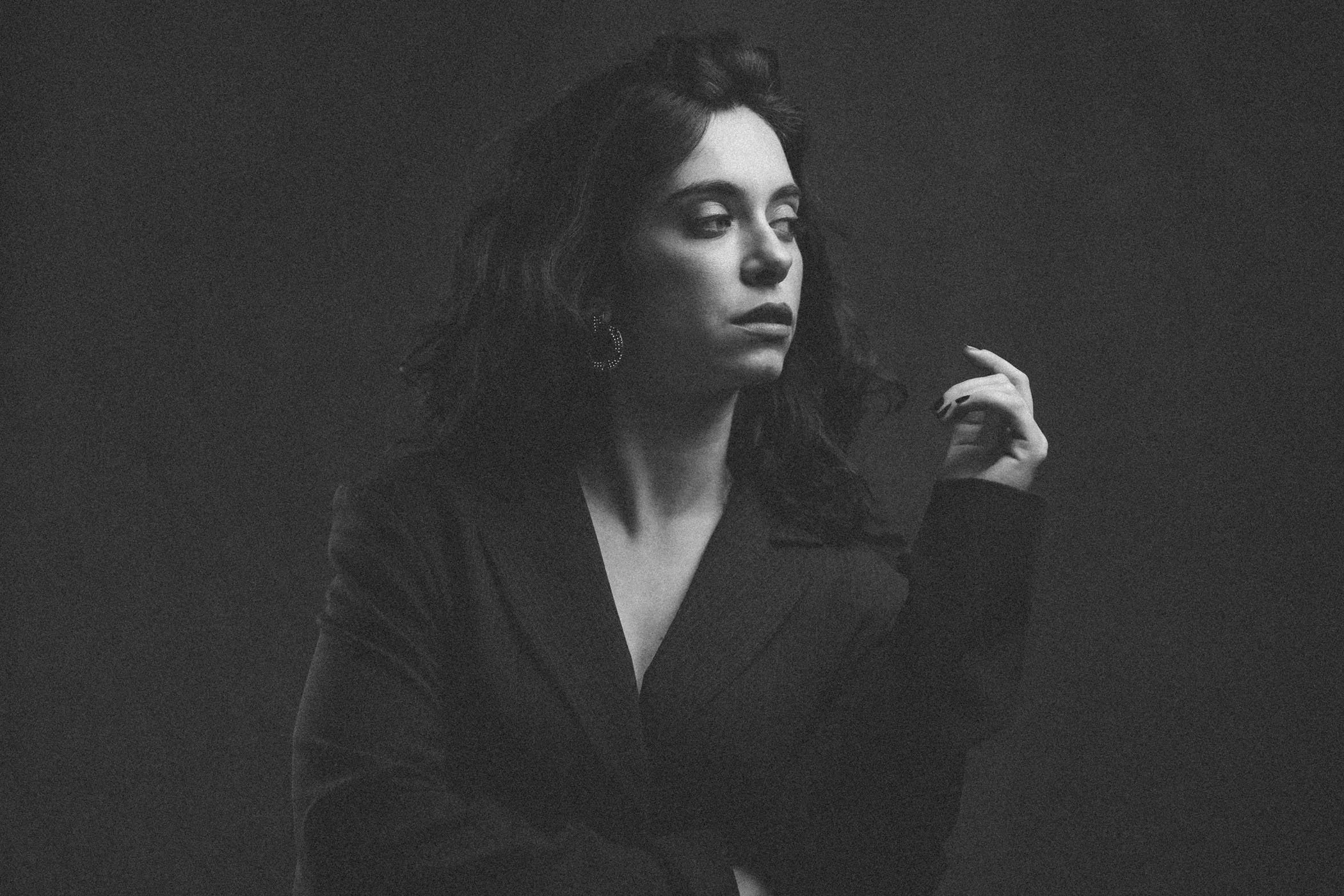 Retrato en blanco y negro de una mujer con cabello rizado, usando aretes grandes y un blazer, con expresión pensativa, en fondo oscuro.
