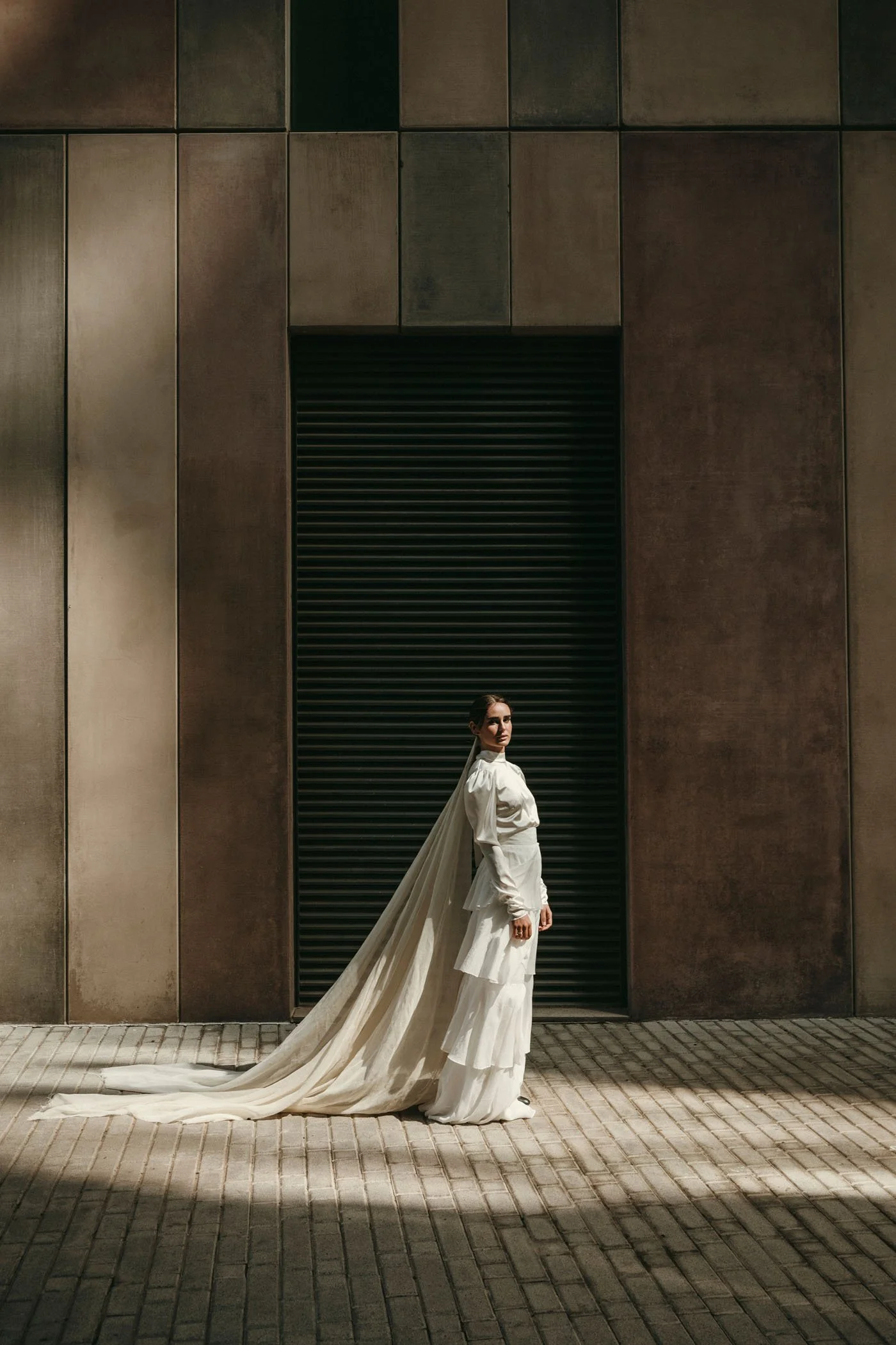 Mujer vestida con atuendo blanco, con capa larga, posando de lado frente a un fondo de pared de metal oscuro y gradas de piedra en la calle.