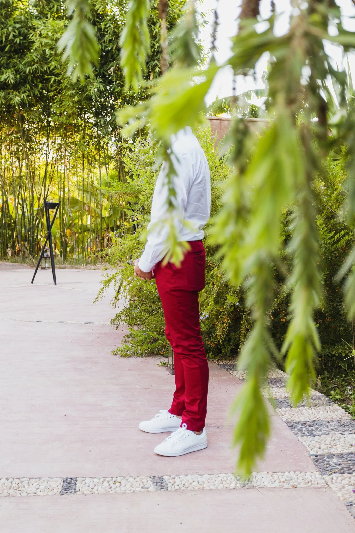 Una persona con pantalón rojo y camiseta blanca de pie en un jardín, vista de lado, rodeada de vegetación, con ramas y hojas en primer plano y fondo de árboles y plantas.