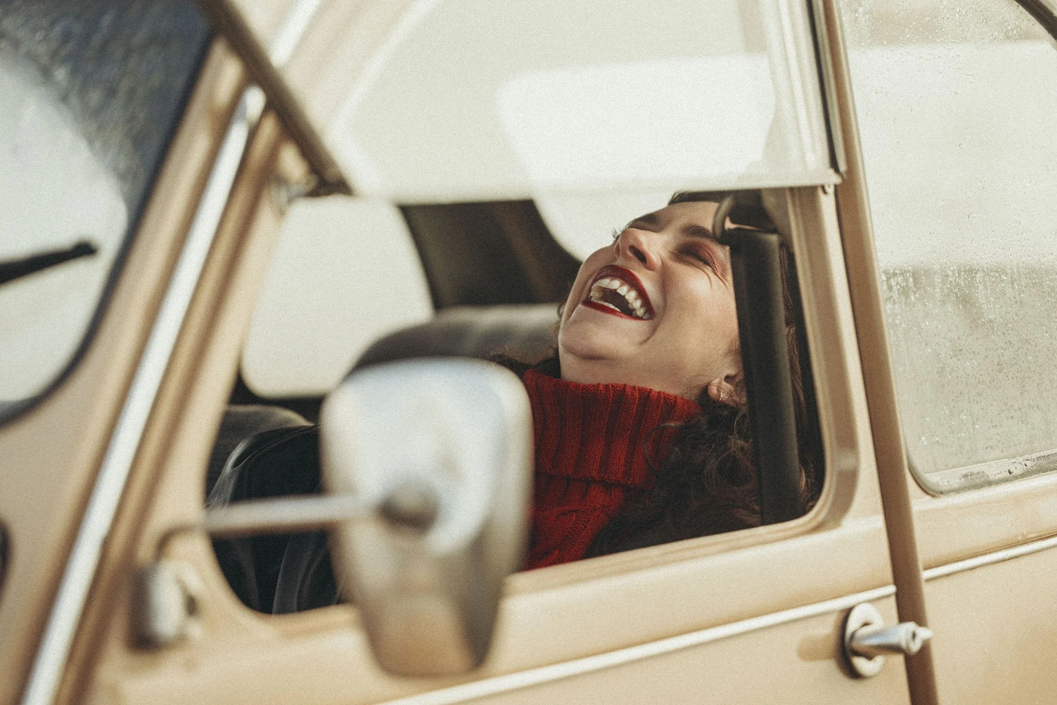 Mujer riendo con la cabeza recostada en el asiento de un coche clásico visto desde la ventana