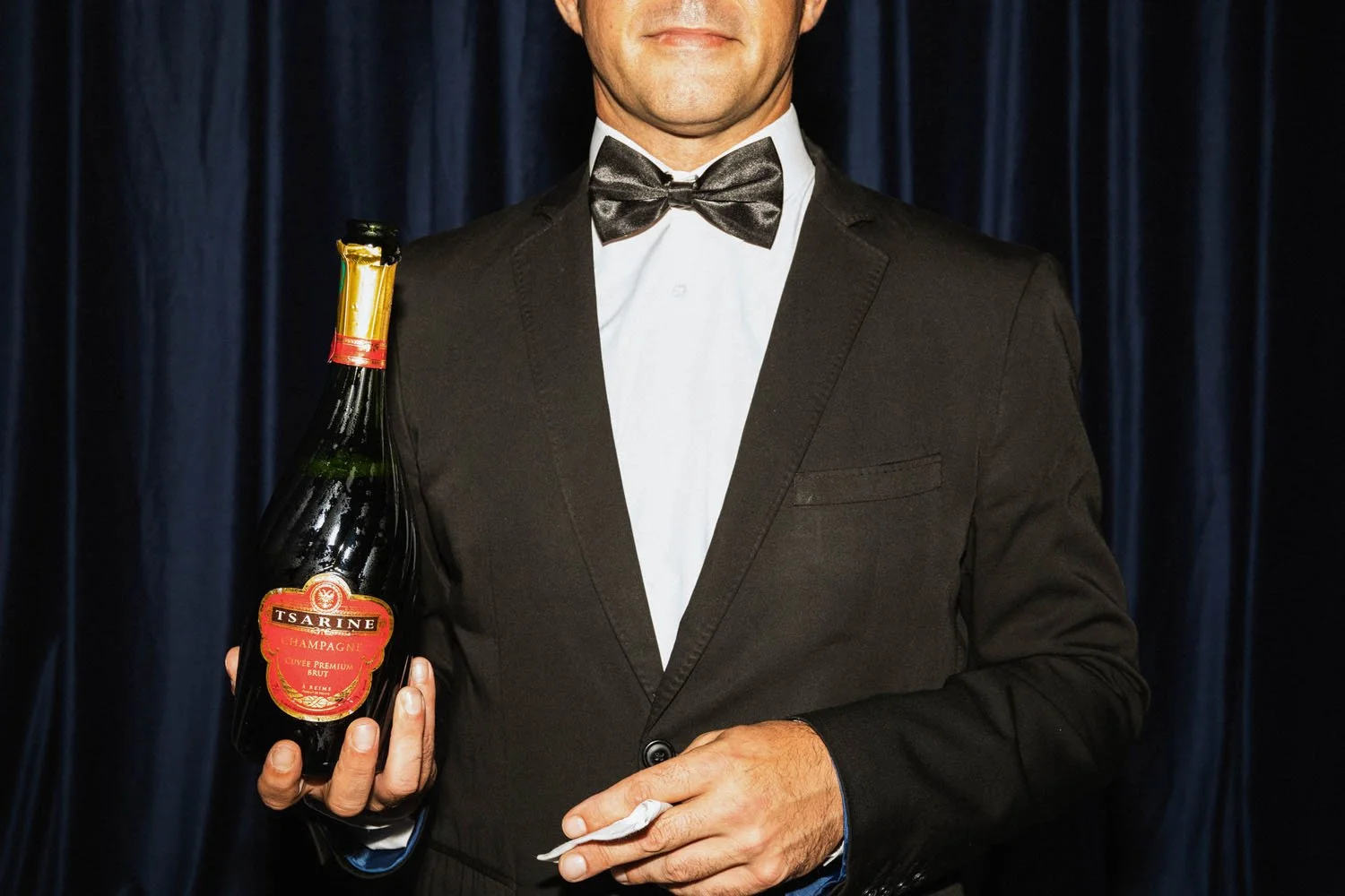 Hombre vestido con traje y moño negro, sosteniendo una botella de champán con etiqueta roja, frente a cortinas azules.